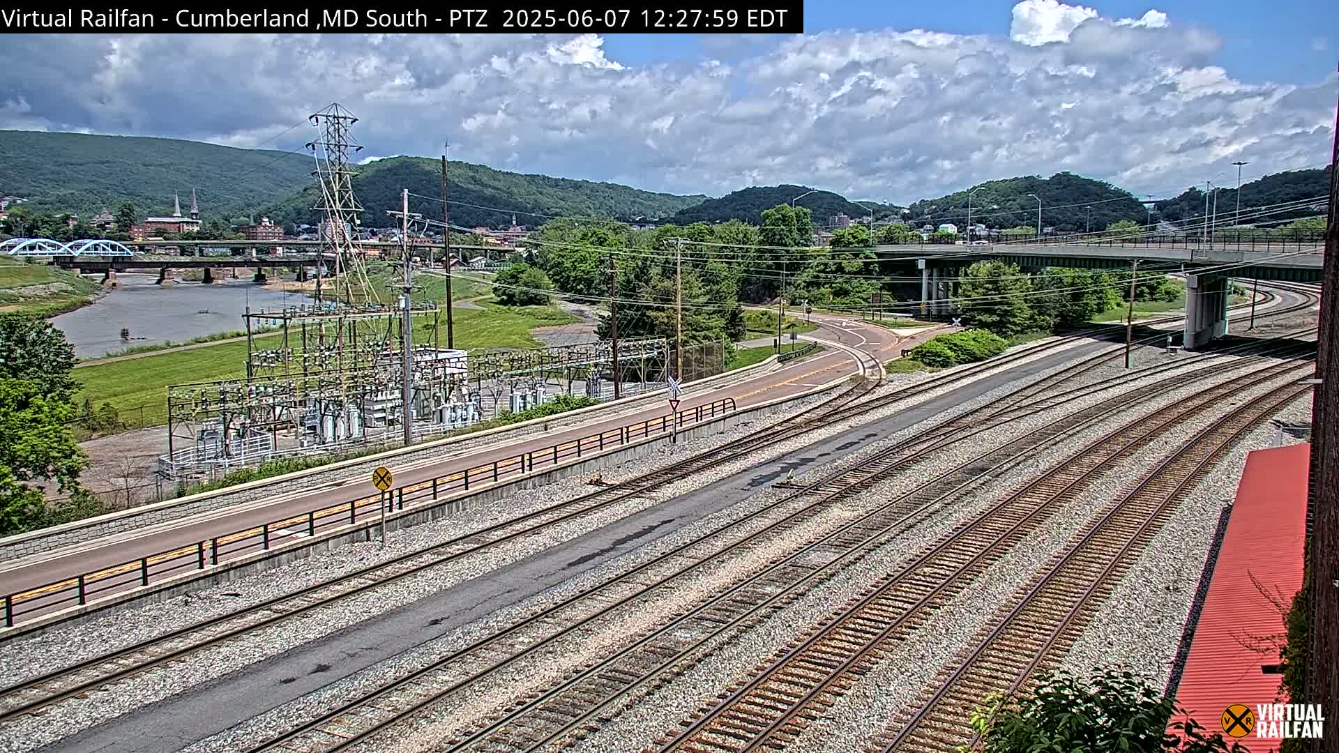 A partly cloudy day reveals a view of railroad tracks, a river, a bridge, and a power substation situated near a town nestled in a valley surrounded by mountains.