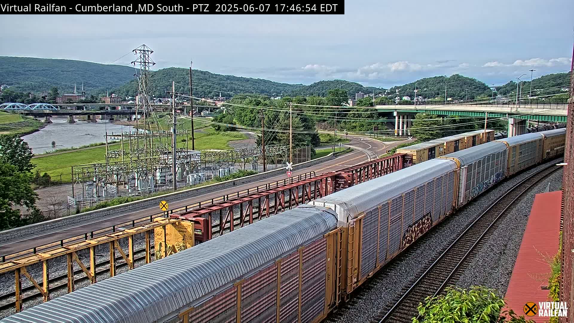 A long freight train curves along railroad tracks beside a river, under a partly cloudy sky, with a power station and a bridge in the background.