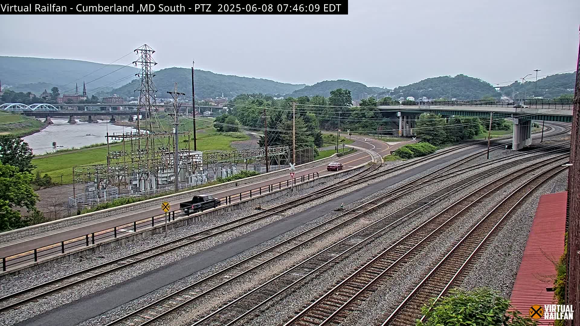 A view of a river, railroad tracks, a road, and a power substation under an overcast sky, with hills and a town in the background.