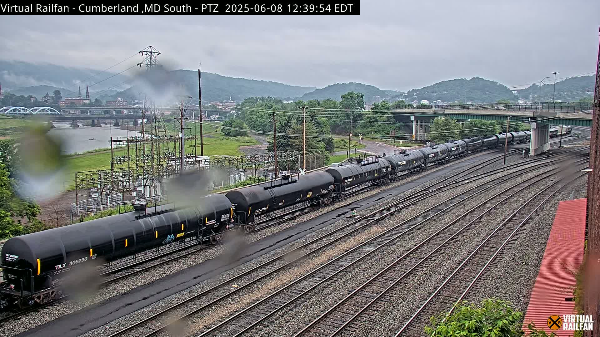 A long freight train of black tanker cars is on railway tracks under an overcast sky, with a river and town visible in the distance.