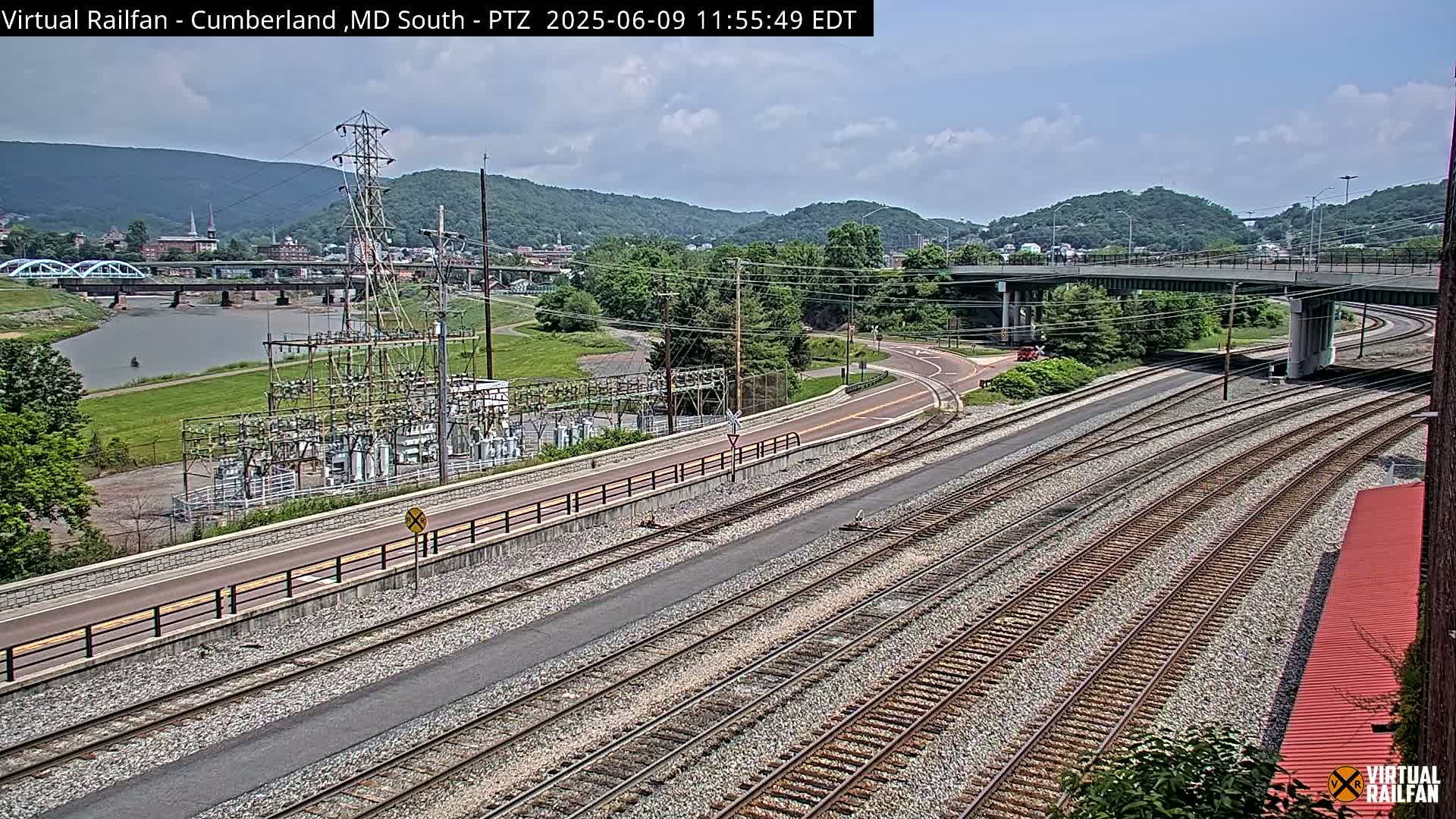 A high-angle view on a partly cloudy day shows several sets of train tracks curving alongside a road and river, with a power substation and a bridge in the distance.