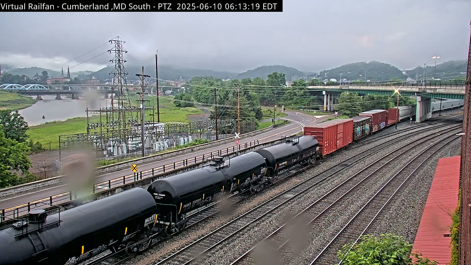 A freight train of black tanker cars and red boxcars travels along railroad tracks under an overcast sky, with a river and a town visible in the background.