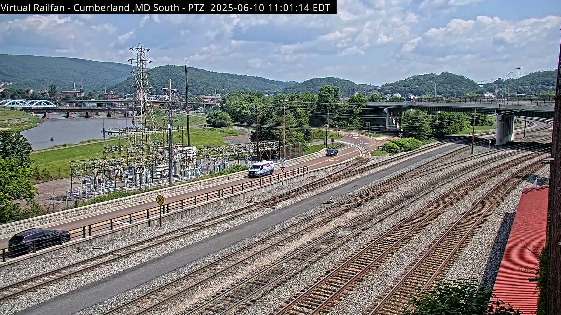 A mostly sunny day reveals a river, a bridge, railroad tracks, a power substation, and a road curving under a highway overpass, all set against a backdrop of green hills.