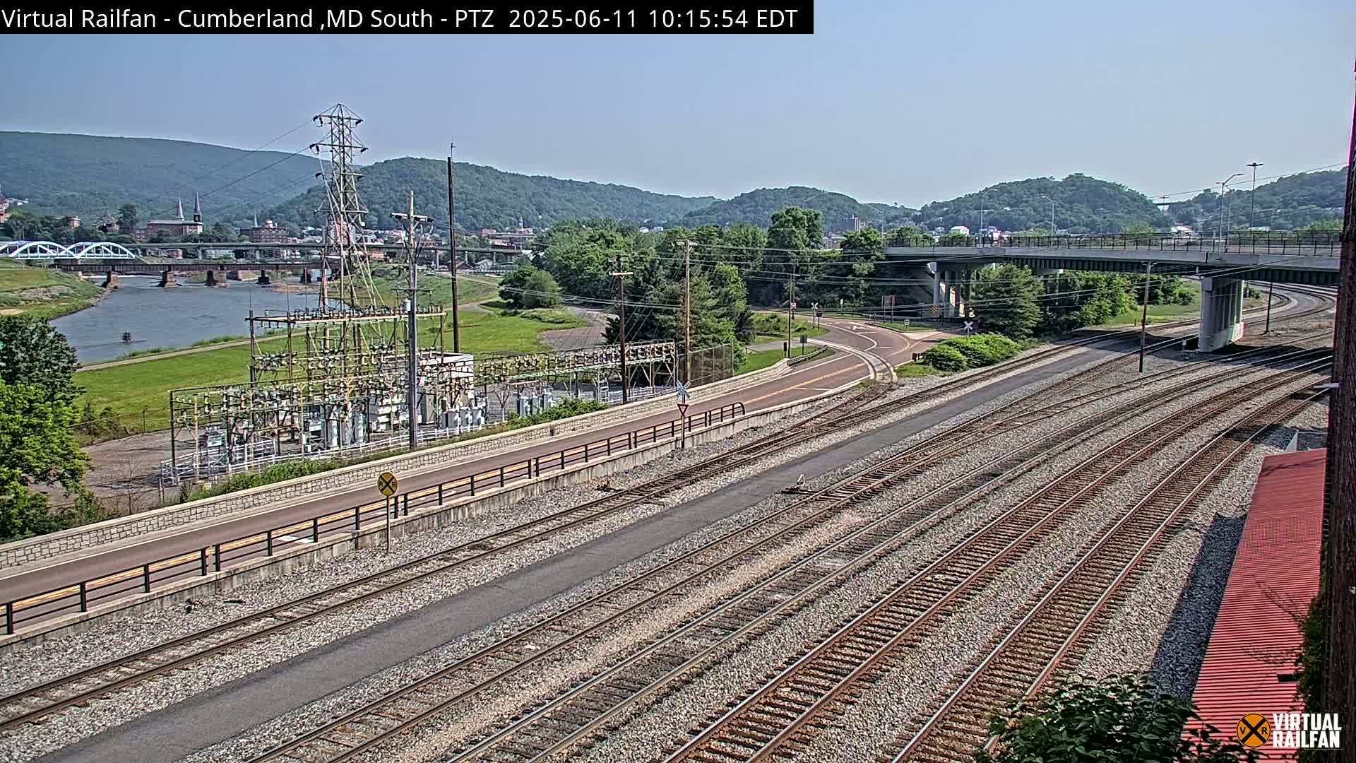 A high-angle view of a train track system next to a river, under a clear sky, with a town and hills in the background.