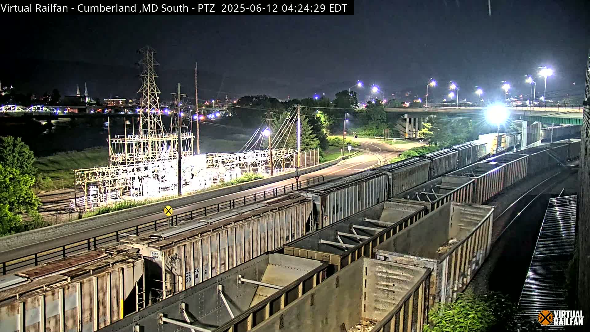 A long freight train is parked at night near an electrical substation beside a river and road, under partly cloudy skies.