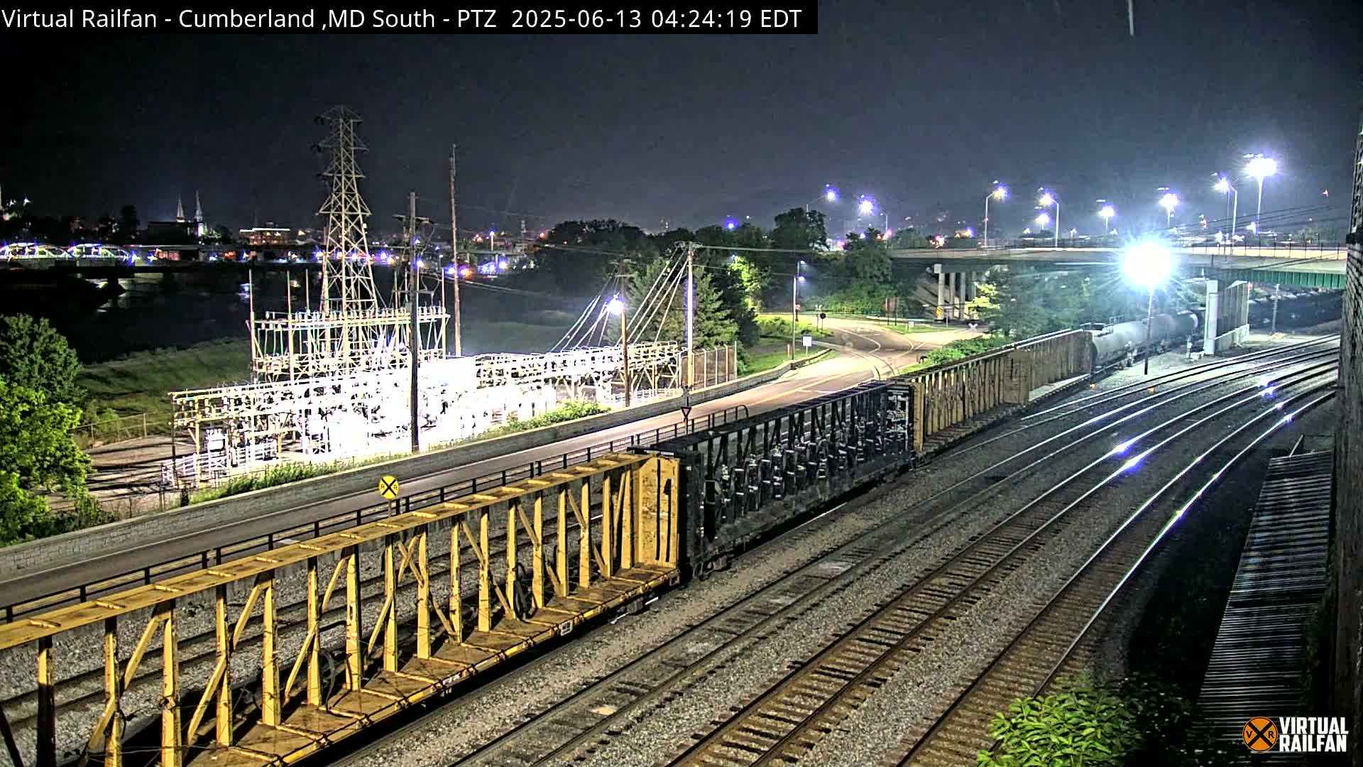 A nighttime view of railroad tracks with several train cars, an electrical substation, a road, and a river in the background under clear skies.