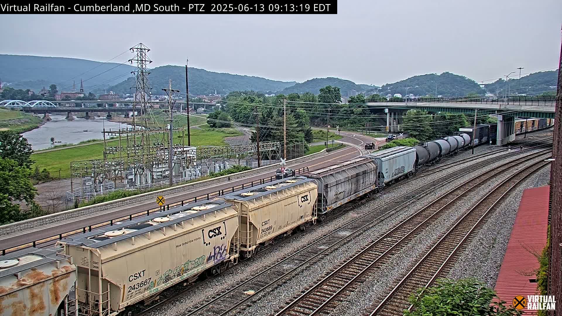 A long freight train is traveling on a set of railroad tracks on an overcast day, passing by a river, bridge, and power station situated against a backdrop of hills.