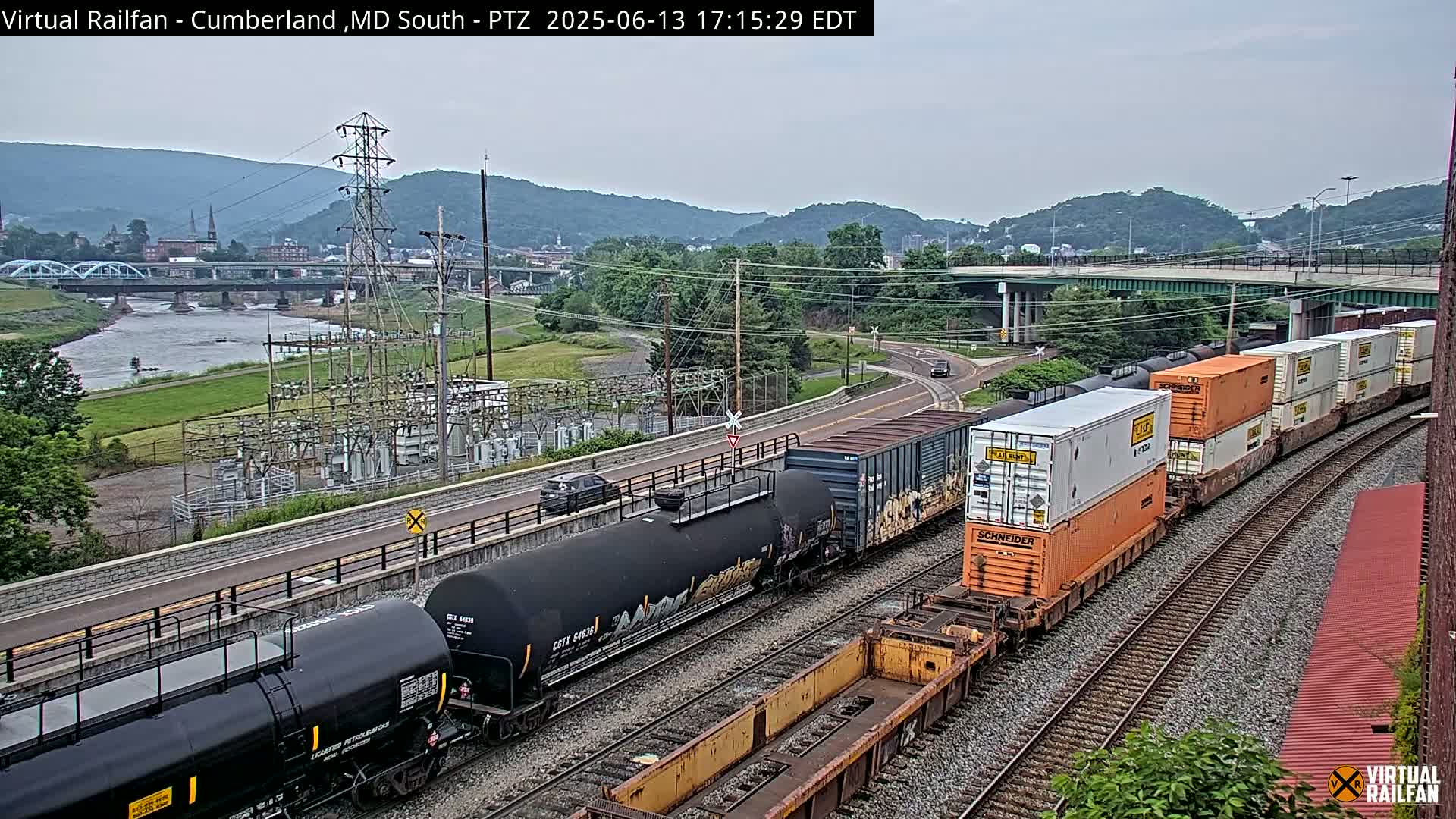 A long freight train, carrying various cargo including tanker cars and shipping containers, is seen traversing a railroad track beside a river, under an overcast sky, with a town and hills in the background.