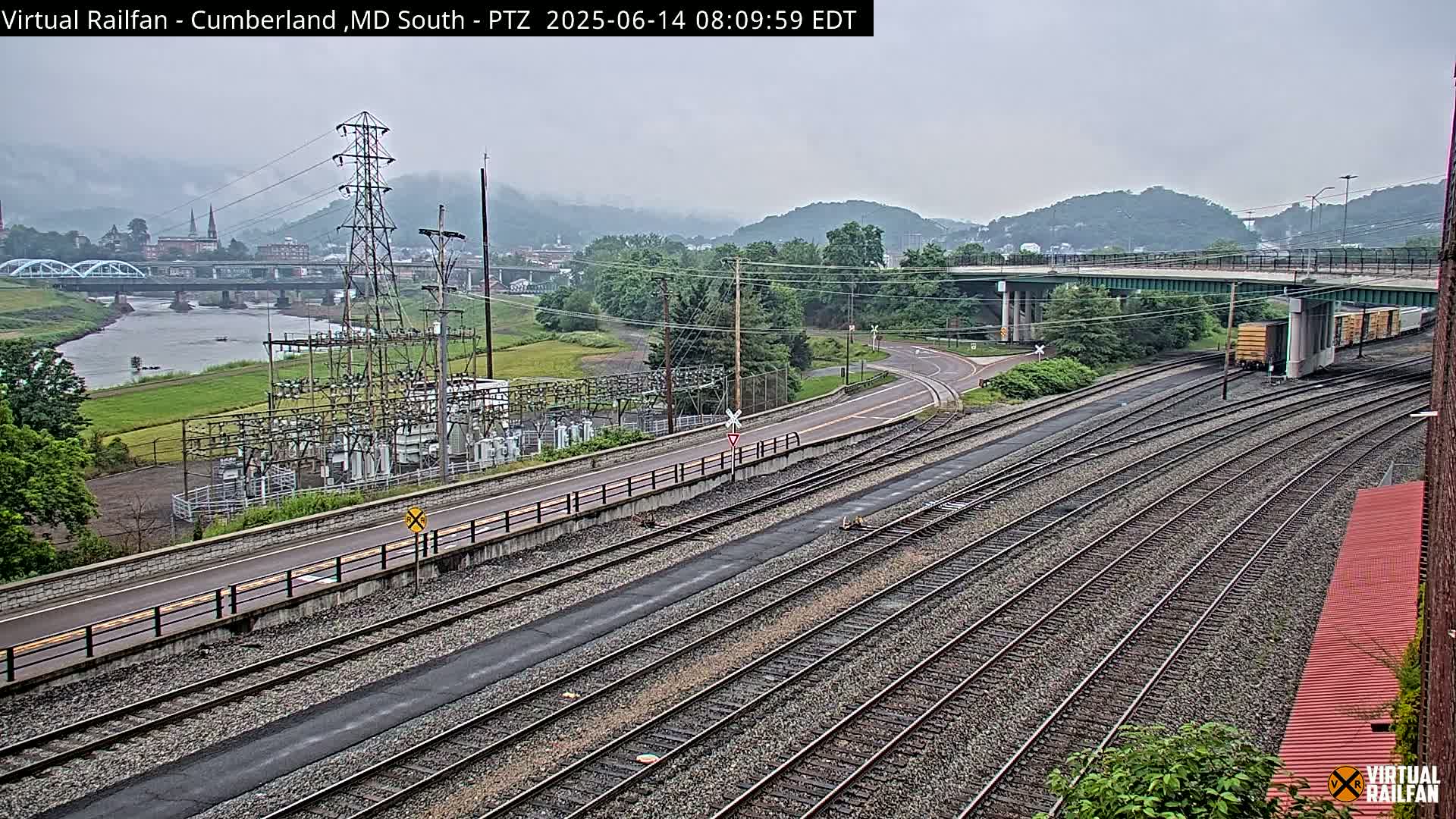 A view of railroad tracks, a road, and a river under an overcast sky, with a power substation and a town in the distance.
