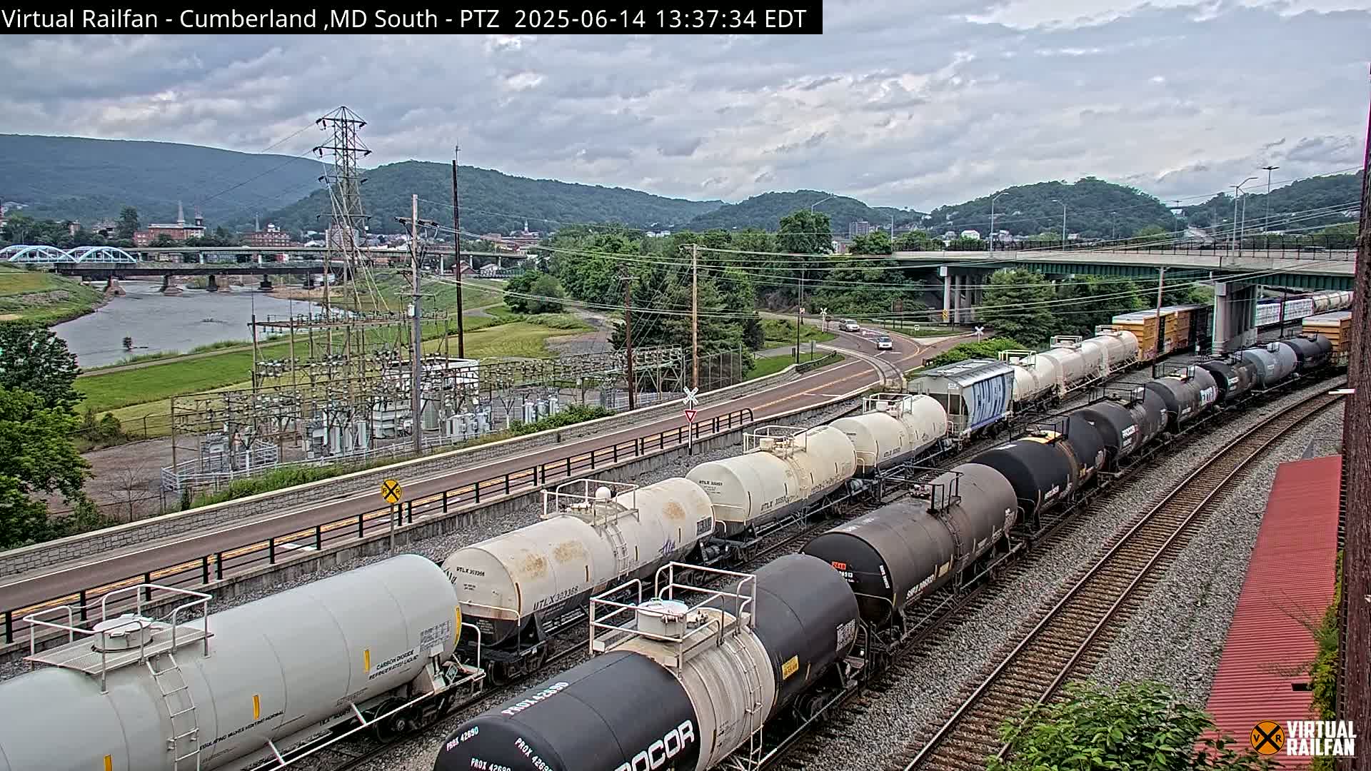 A long freight train of tanker cars curves along a set of railroad tracks under an overcast sky, with a river, bridge, and power substation visible in the background.