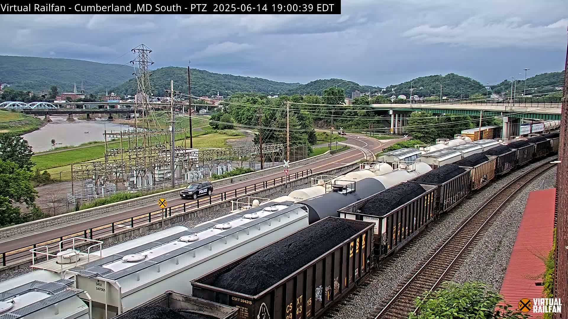 A long freight train of tanker cars curves along a set of railroad tracks under an overcast sky, with a river, bridge, and power substation visible in the background.
