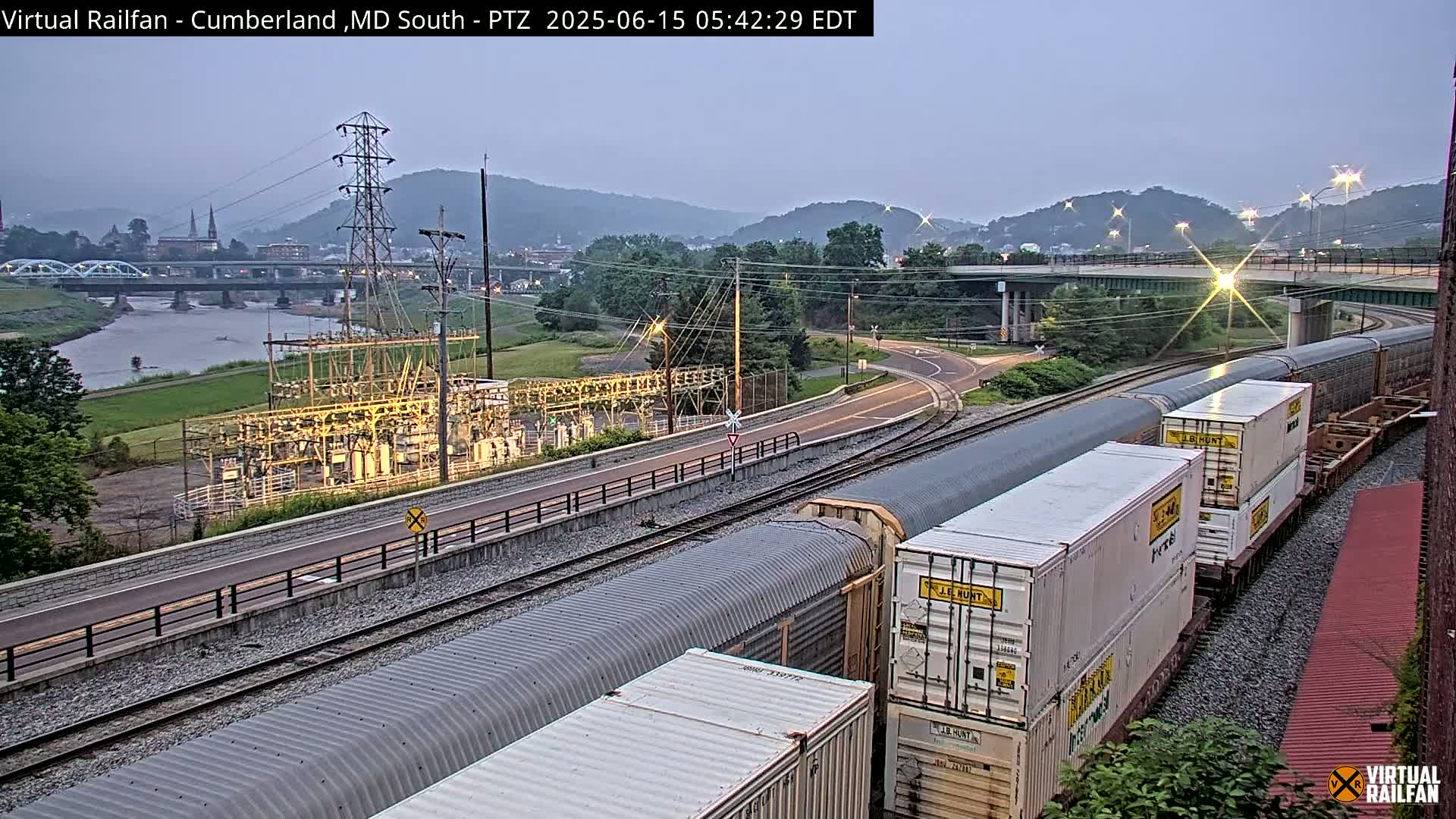 A long freight train sits on railroad tracks beside a river, under a dusky sky, with a power station and a bridge in the background.