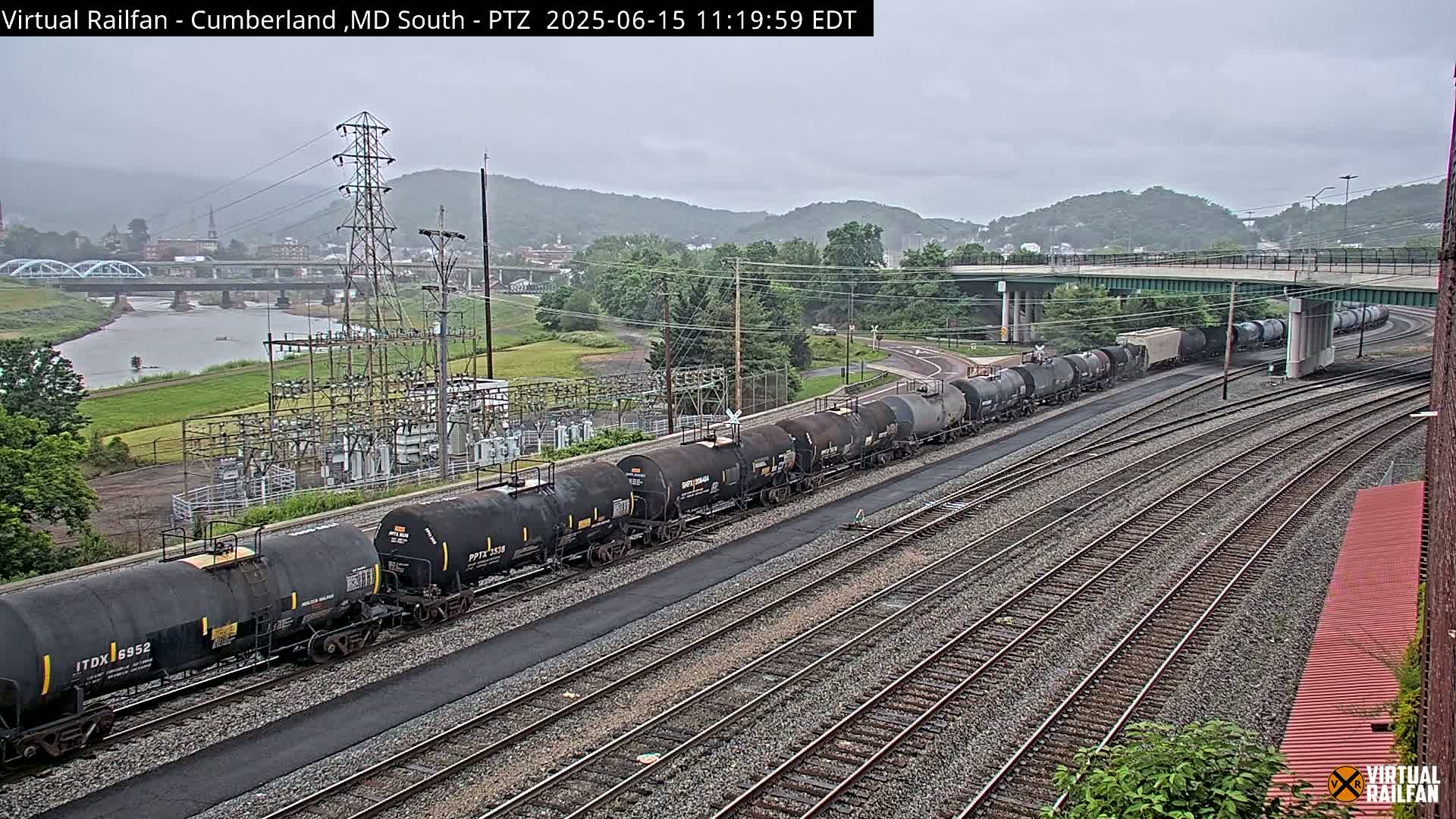 A long freight train of tanker cars sits on a set of railroad tracks on an overcast day, with a river, bridge, and power station visible in the background.