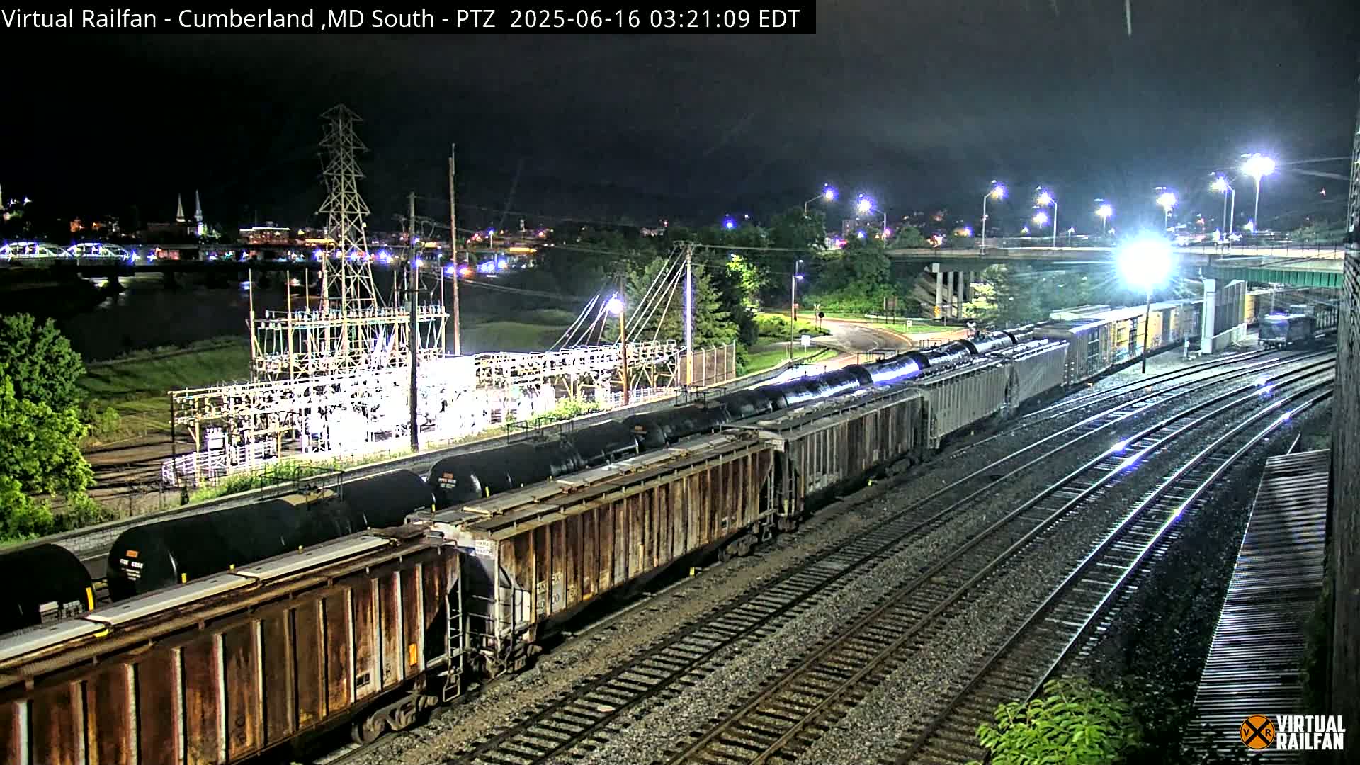 A long freight train of tank cars and boxcars is visible at night, passing near an electrical substation and under a highway overpass.