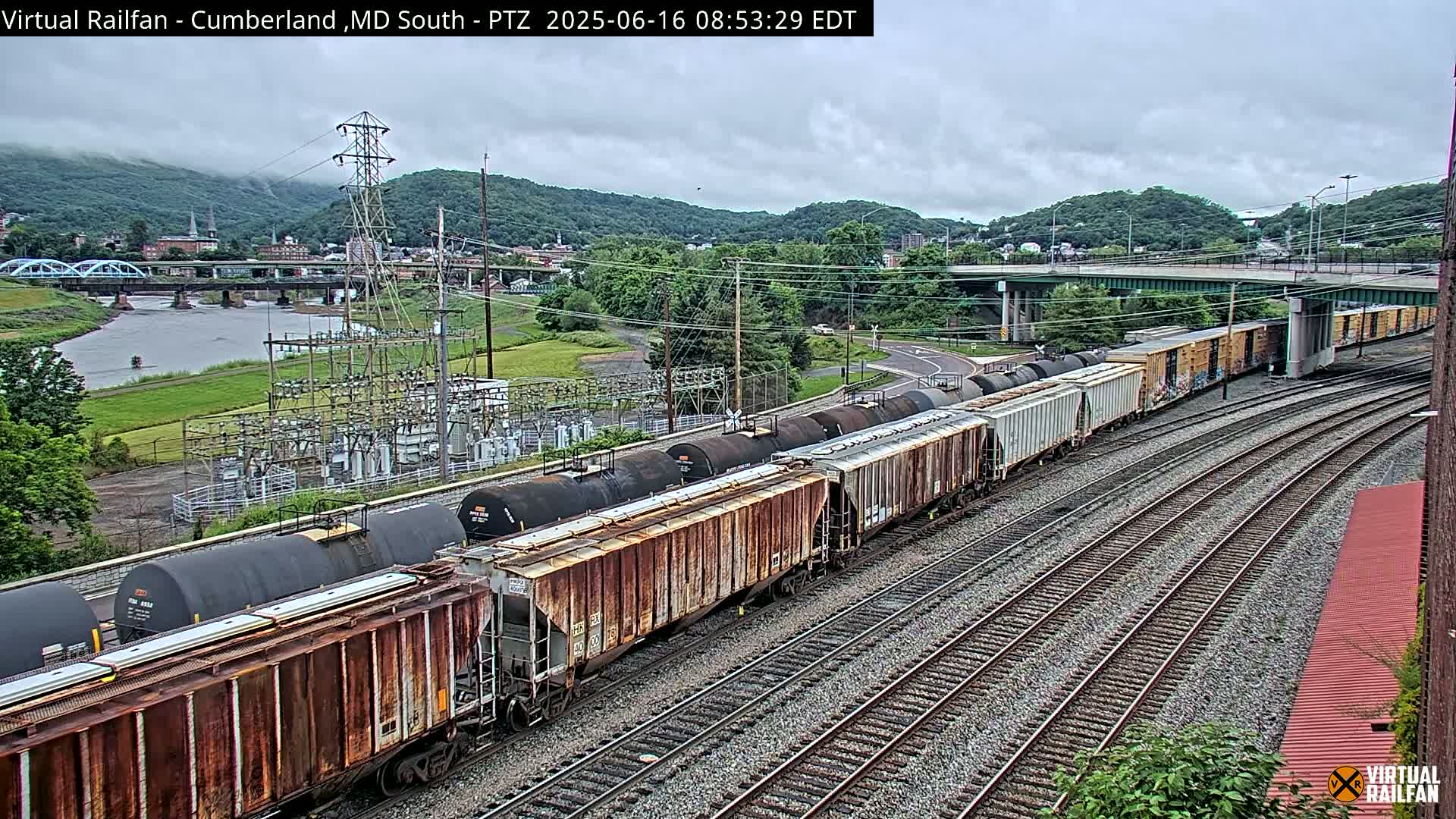 A long freight train, consisting of rusty boxcars and tanker cars, sits on multiple train tracks next to a river under an overcast sky, with a town and hills in the background.