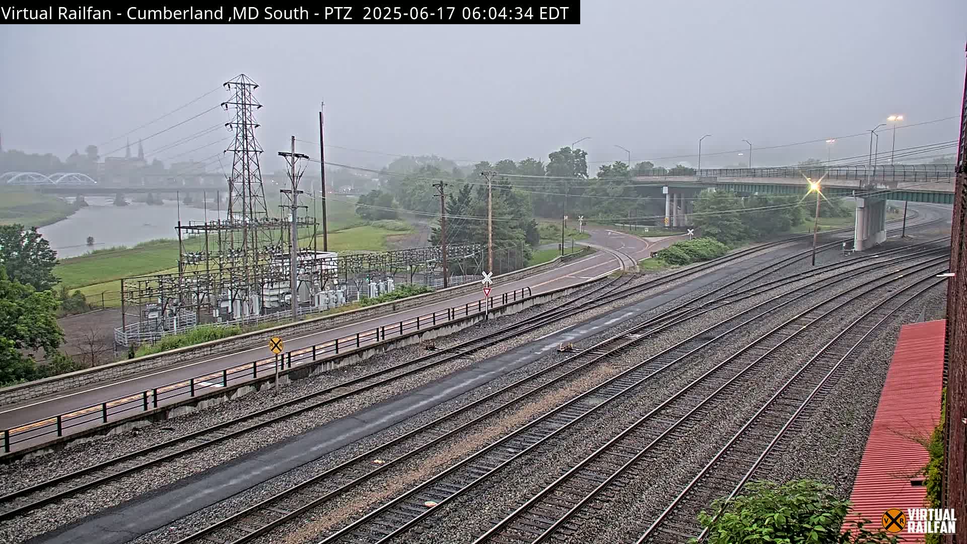 A hazy, overcast day reveals a view of multiple train tracks intersecting with a road near a river and power substation.