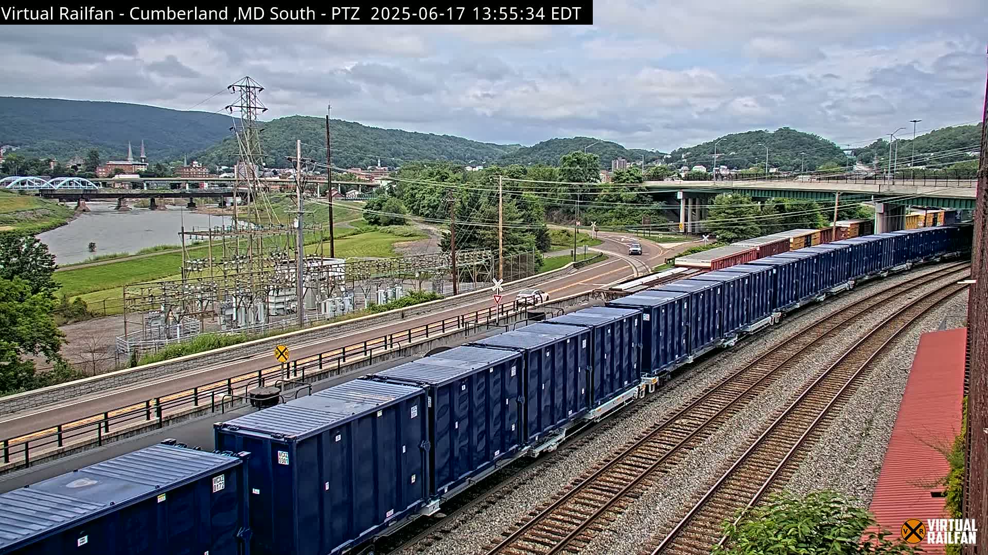 A long freight train of dark blue containers curves along railroad tracks under a cloudy sky, past a river, power plant, and town nestled against hills.