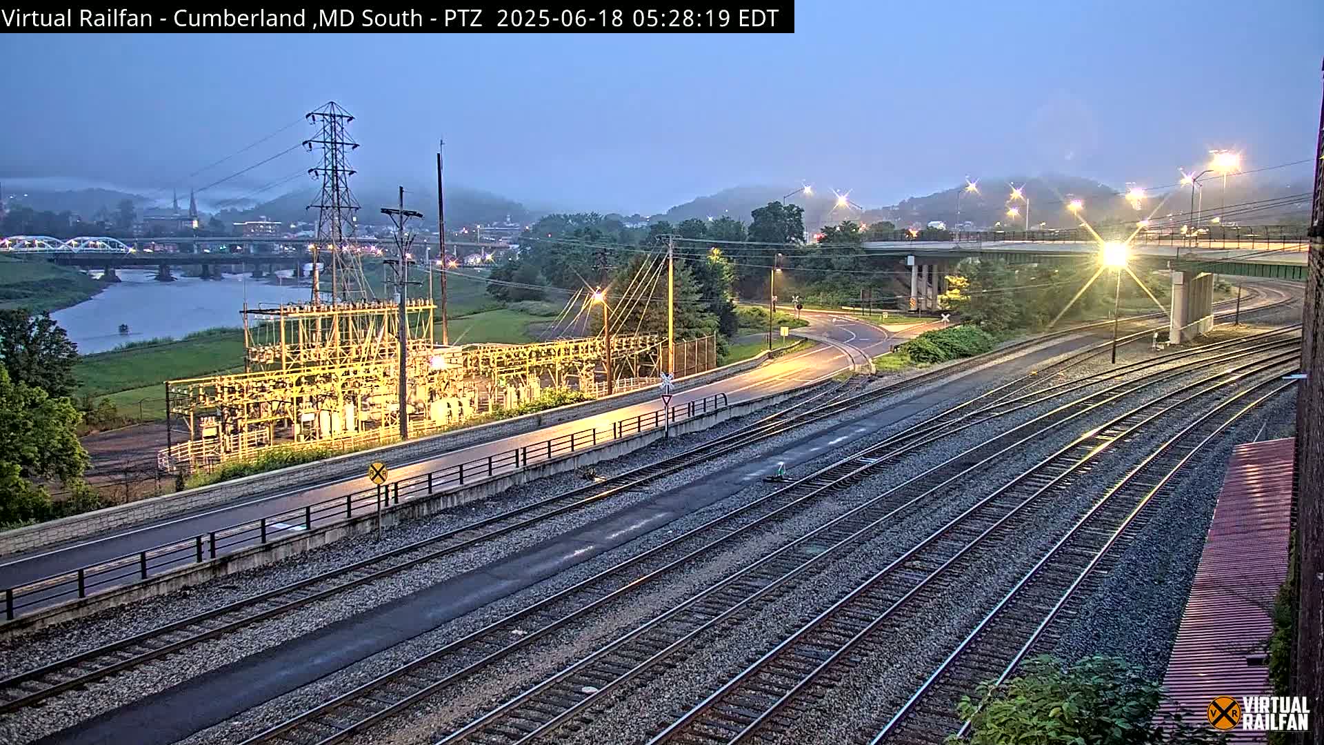 A twilight view of a river, bridges, railroad tracks, a power substation, and roads under a mostly clear sky.