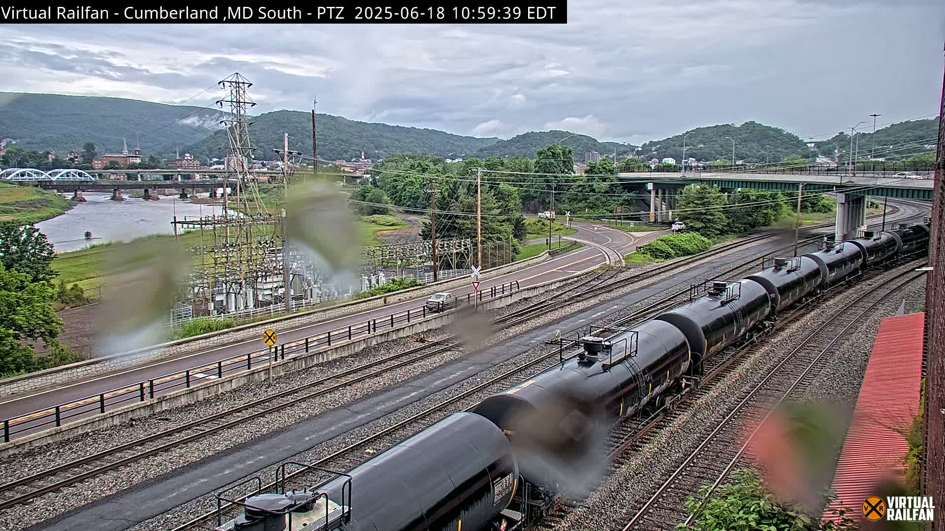 A long freight train of black tanker cars curves along railroad tracks next to a river, under an overcast sky, with a roadway and power station visible in the background.
