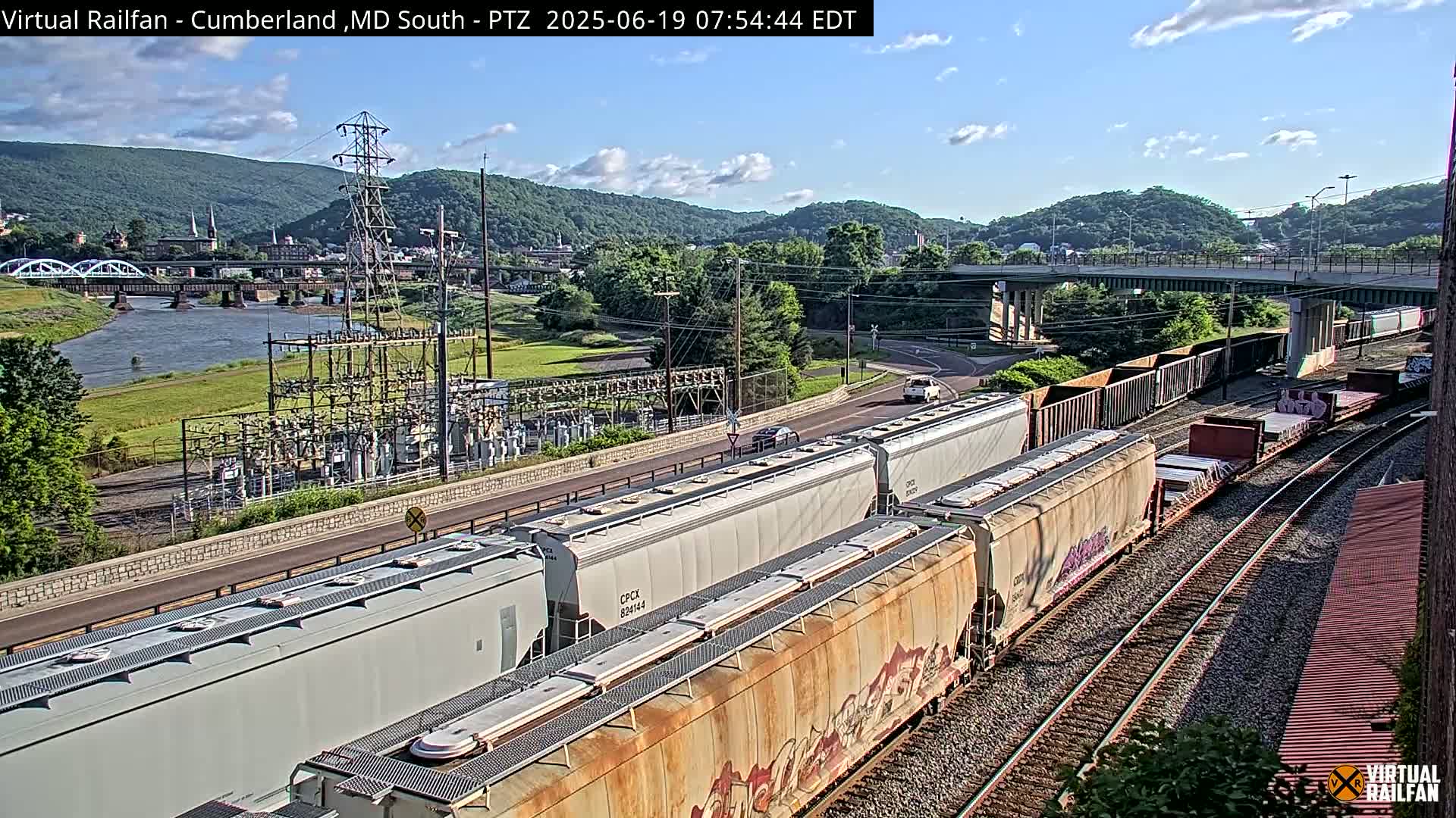 A long freight train sits on railroad tracks next to a river, under a mostly sunny sky, with a power plant and a bridge visible in the background.