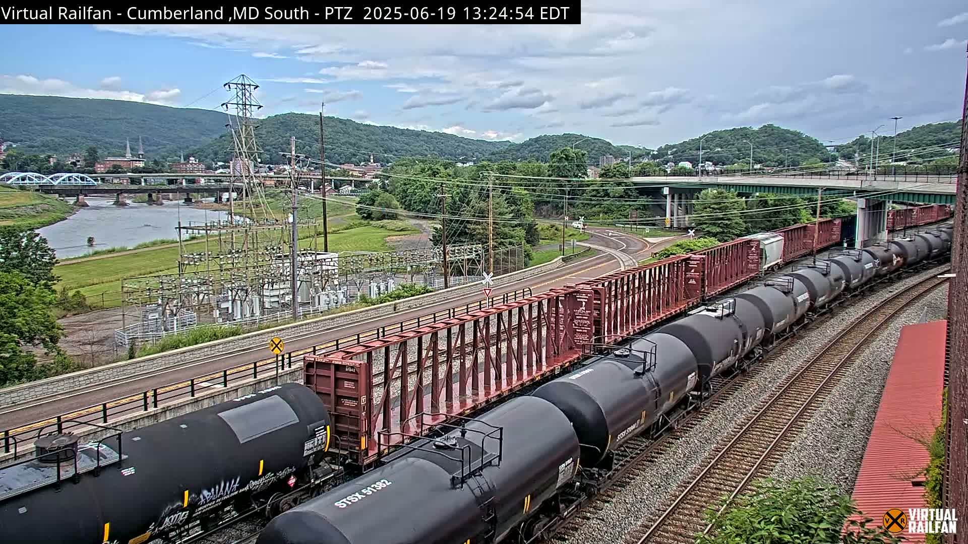 A long freight train of tanker cars and boxcars curves along railroad tracks under a partly cloudy sky, passing by a river, bridge, and power substation near a town nestled in a valley.