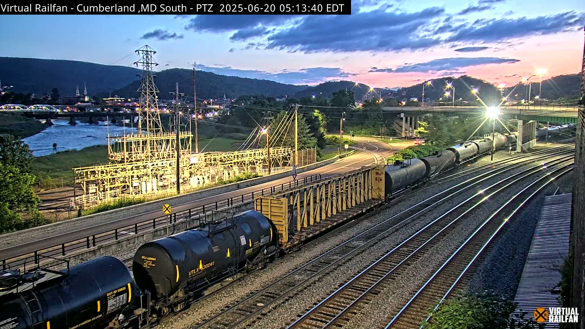 A freight train passes by an electrical substation at dusk near a river and mountains under a partly cloudy sky.