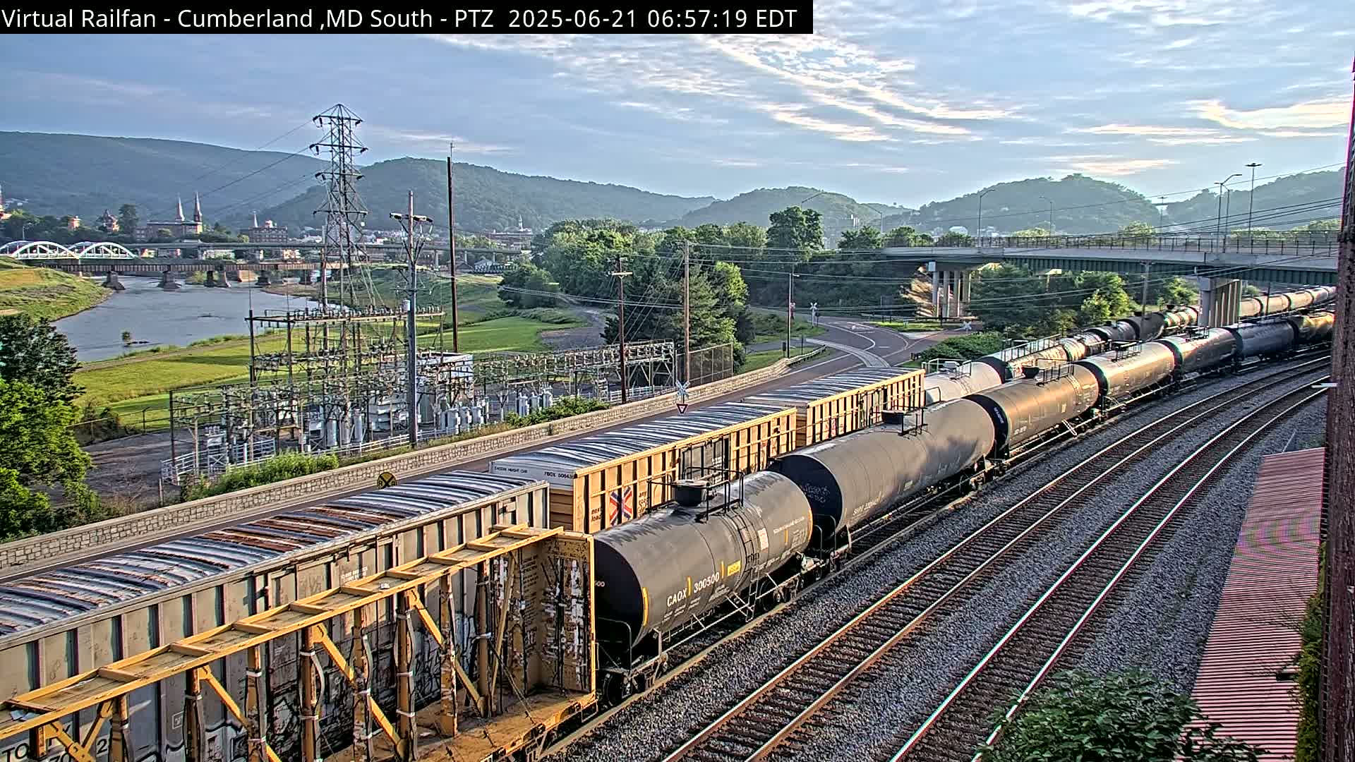 A long freight train of tanker cars and boxcars sits on a set of railroad tracks under a partly cloudy sky, with a river, bridge, and power station visible in the background.