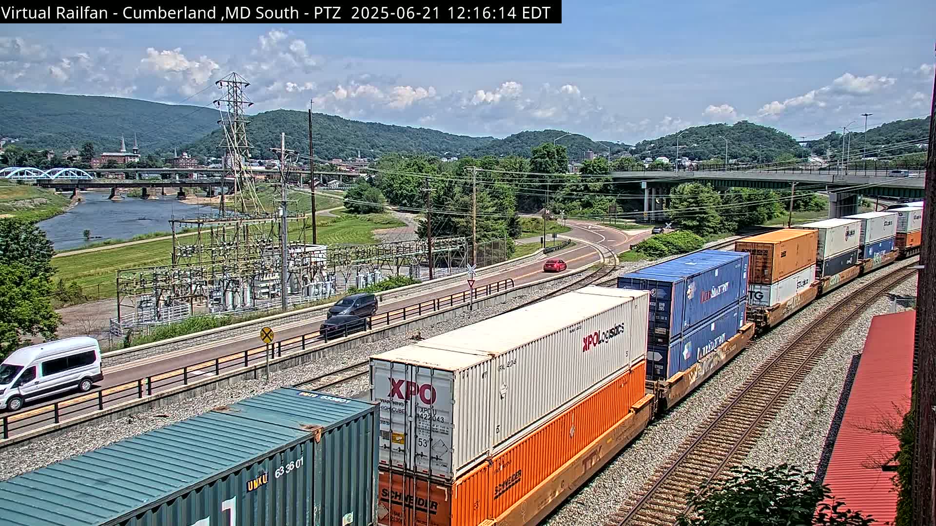 A long freight train carrying multiple shipping containers moves along railroad tracks beside a road and river, under a partly cloudy sky.