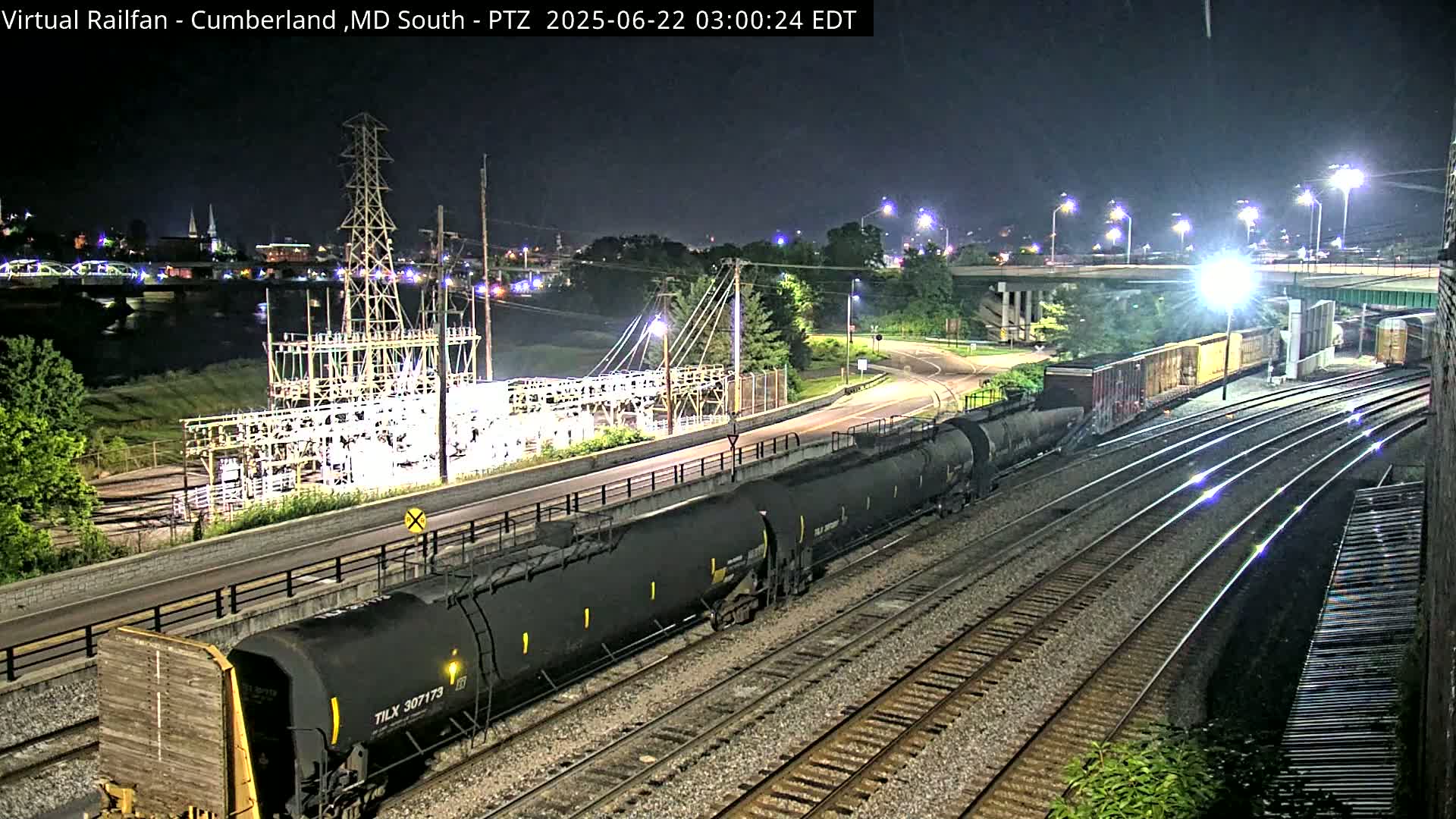 A long freight train passes by a power substation at night, near a river and roadway under bright lights.