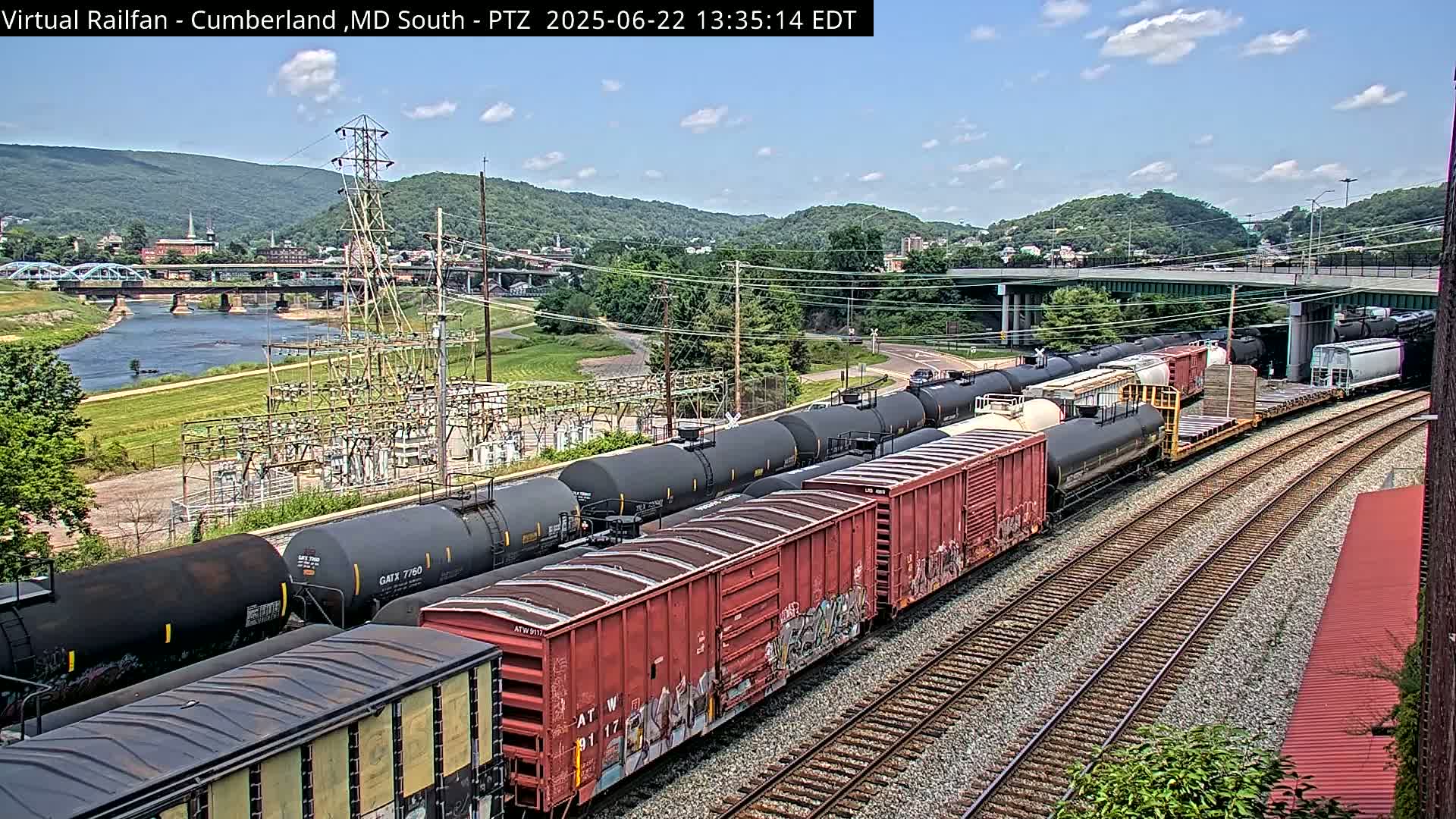 A long freight train carrying tanker cars and boxcars is parked on a railroad track next to a river under a partly cloudy sky, with a town and mountains in the background.