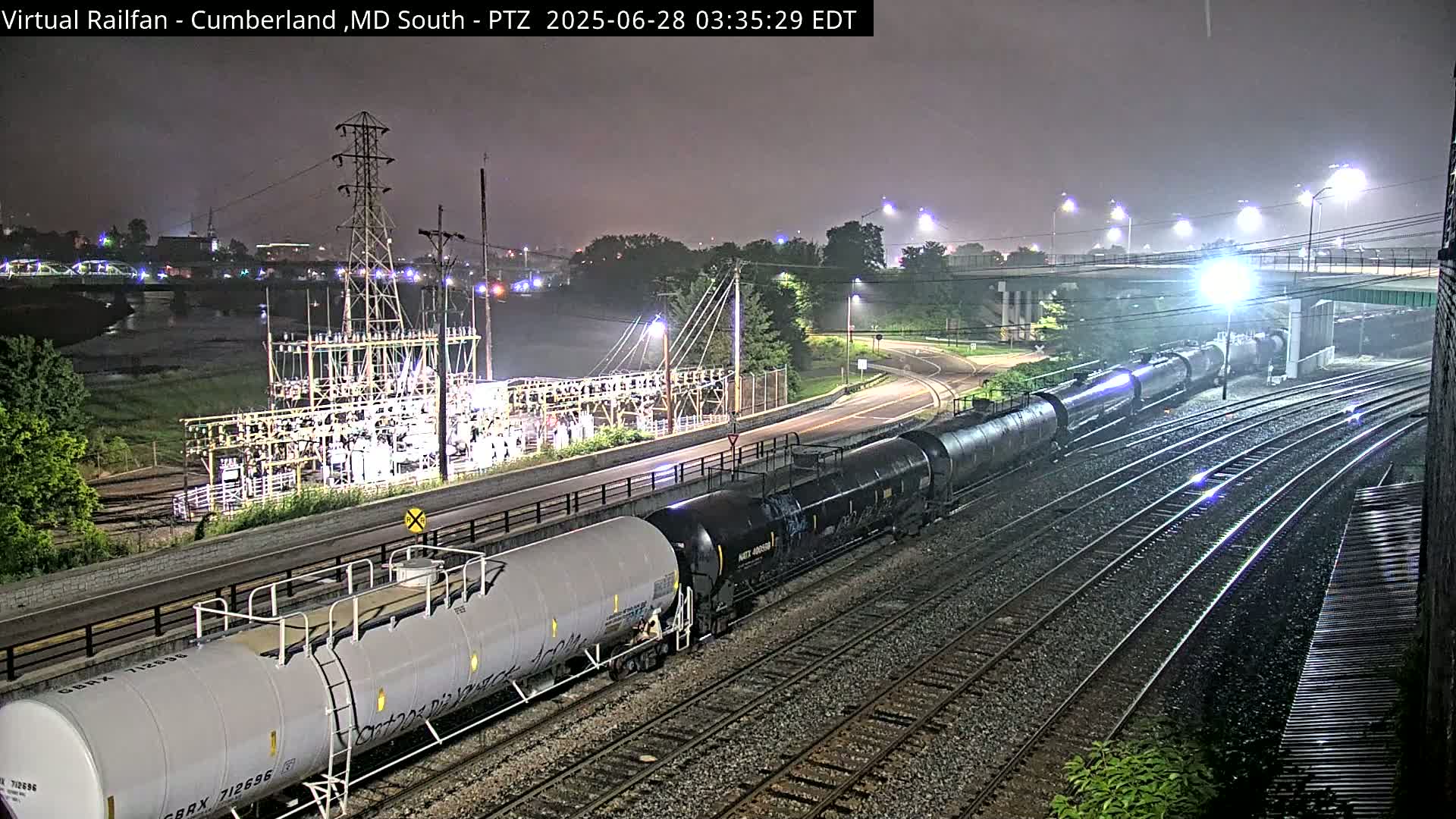At night, under a misty sky, a long freight train of tanker cars moves along railroad tracks near a power substation and a roadway, with a bridge visible in the background.