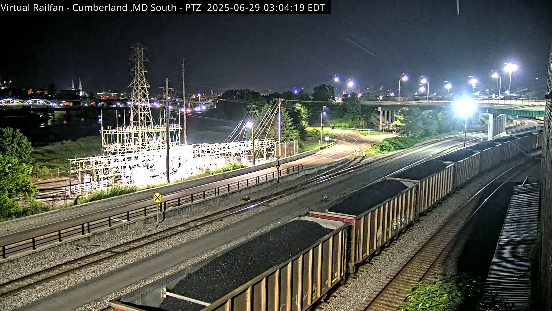 A nighttime view of a train carrying coal along railroad tracks near a river, with an electrical substation and highway overpass in the background.