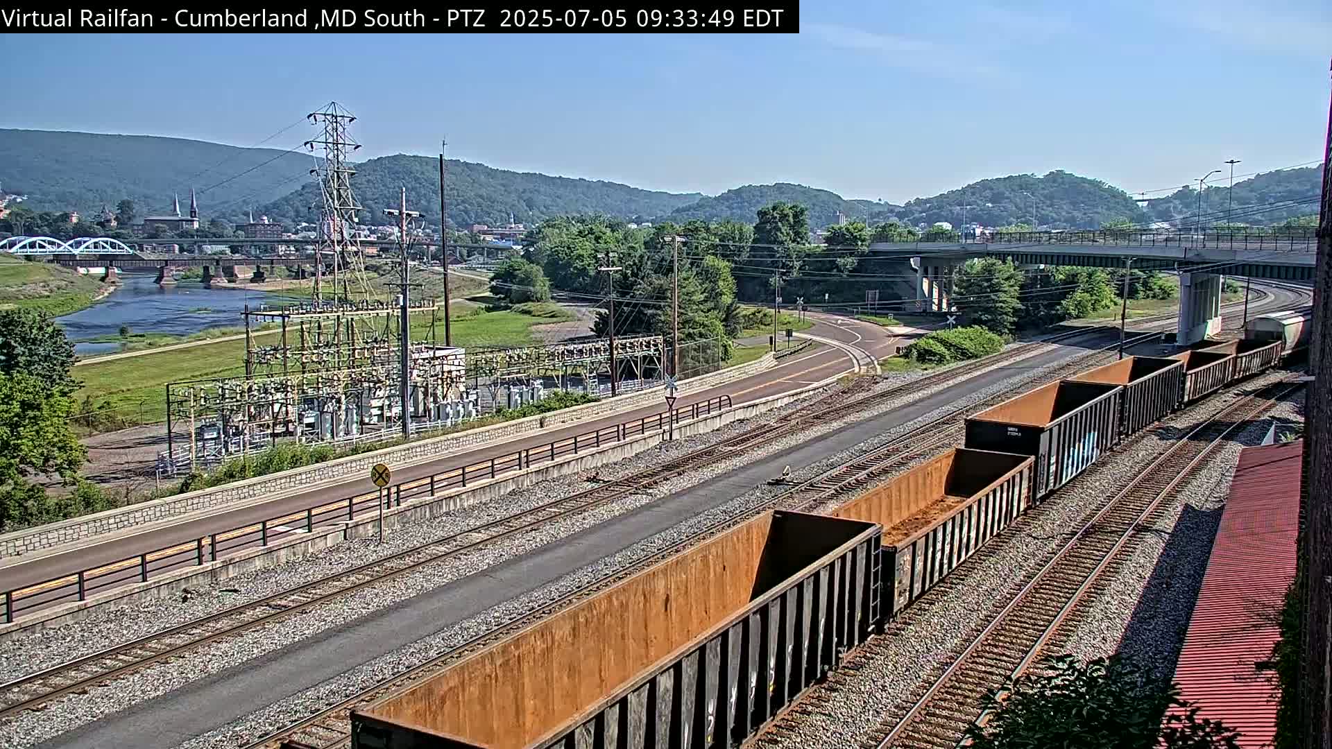 A sunny day reveals a railway scene with several empty train cars on parallel tracks, near a river, power station, and a highway overpass, all set against a backdrop of hills and a small town.