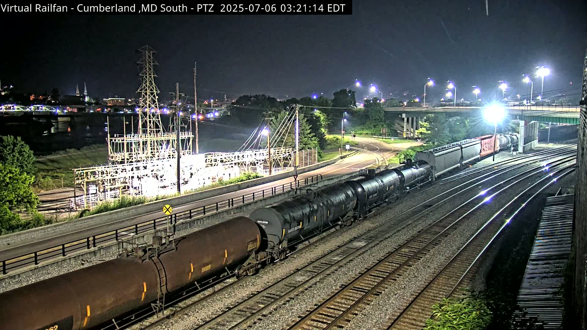 A long freight train of tanker cars passes an electrical substation at night under bright lights.