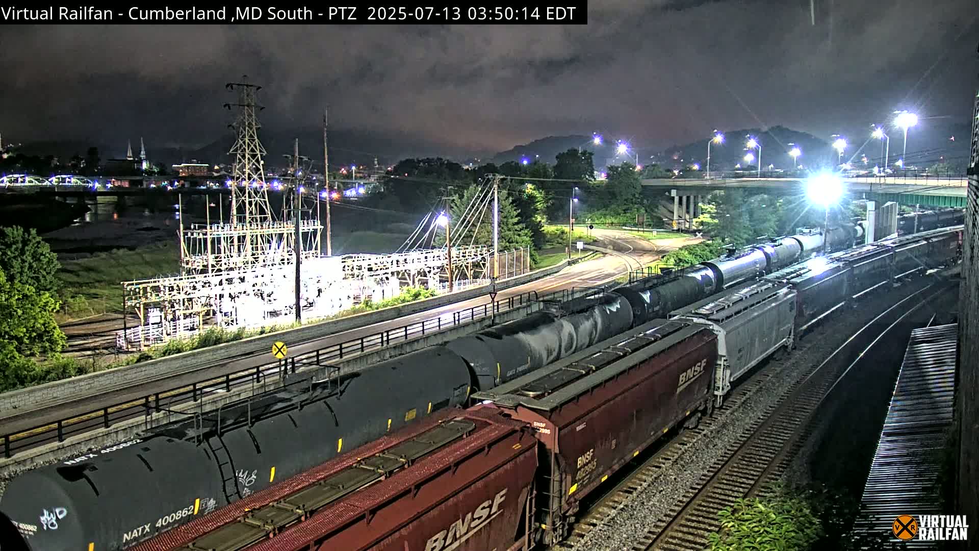 At night, under a partly cloudy sky, a long freight train with numerous tanker cars and boxcars passes by an electrical substation near a roadway and river.