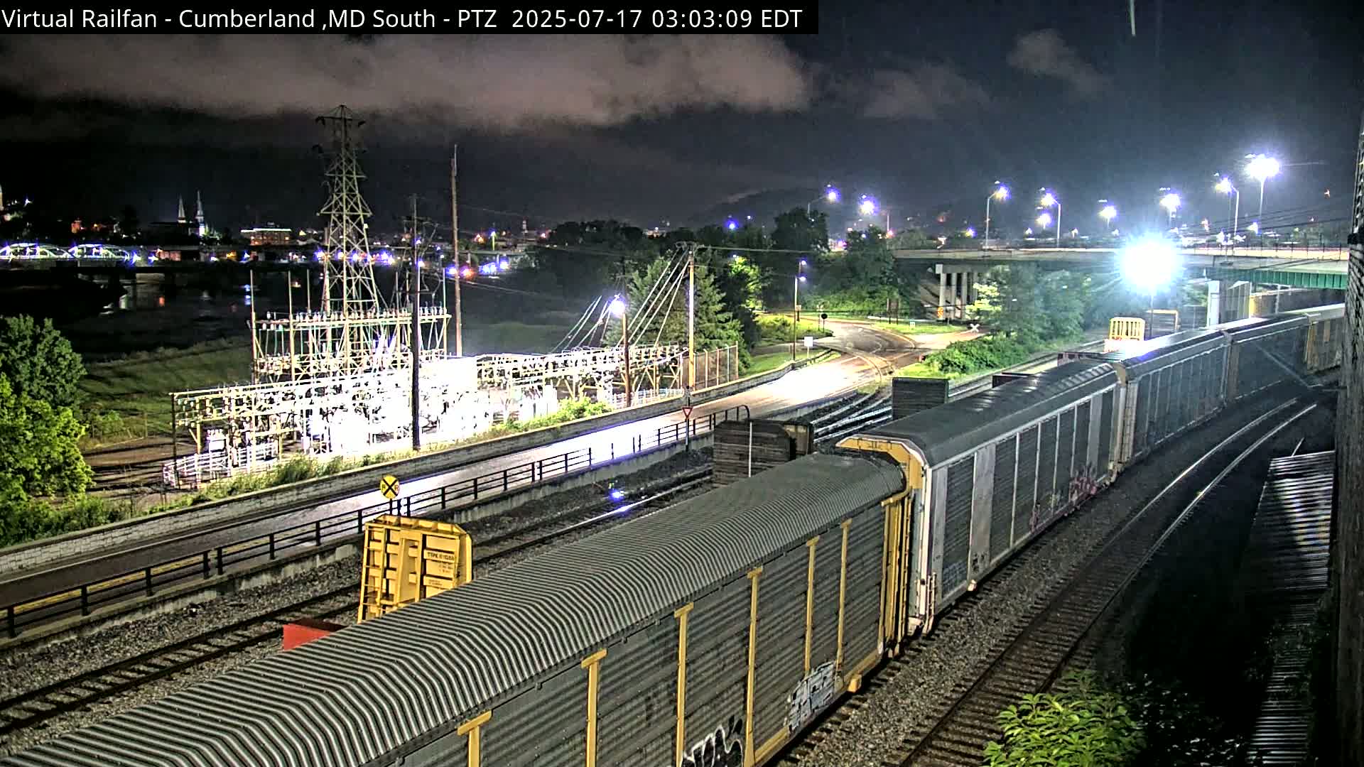 A long freight train travels along railroad tracks at night under cloudy skies, passing an electrical substation and a roadway.