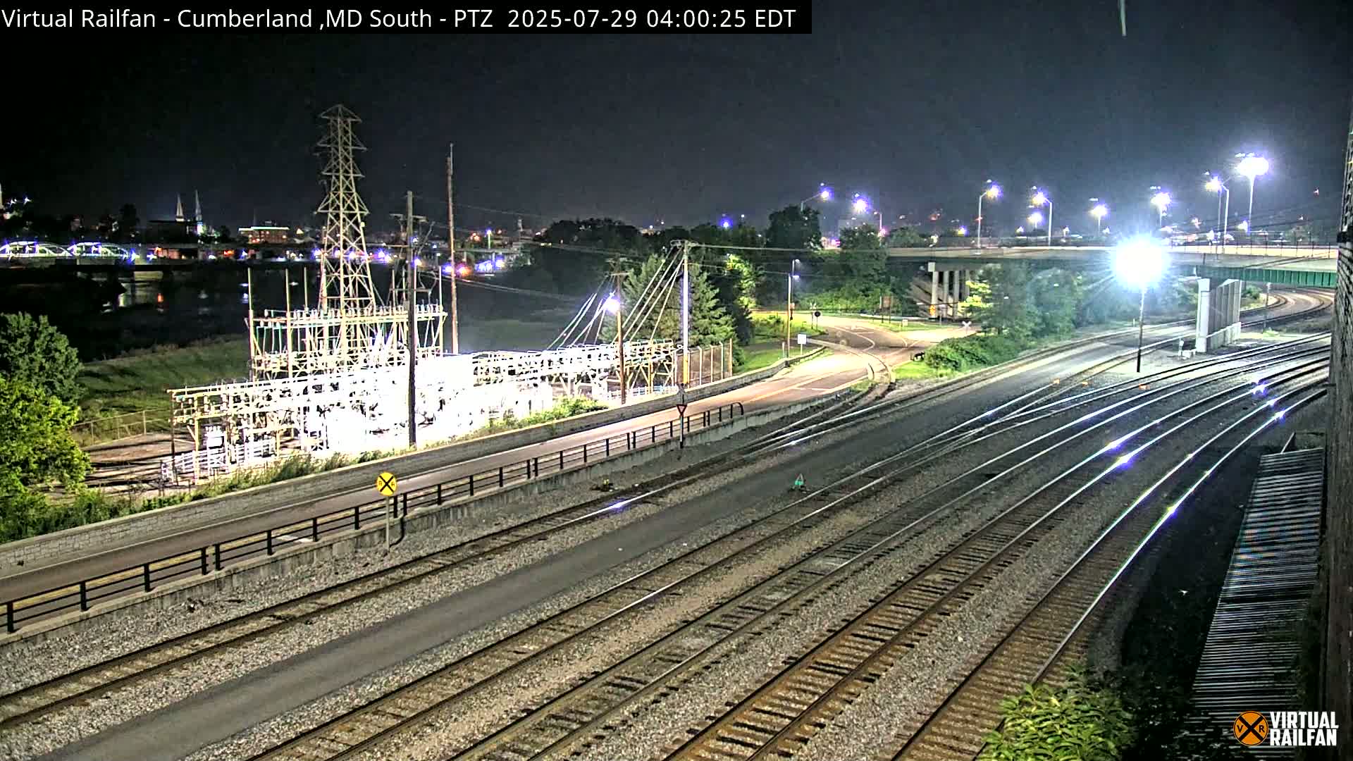 A nighttime view of railroad tracks intersecting with roads and a power substation, under clear skies.