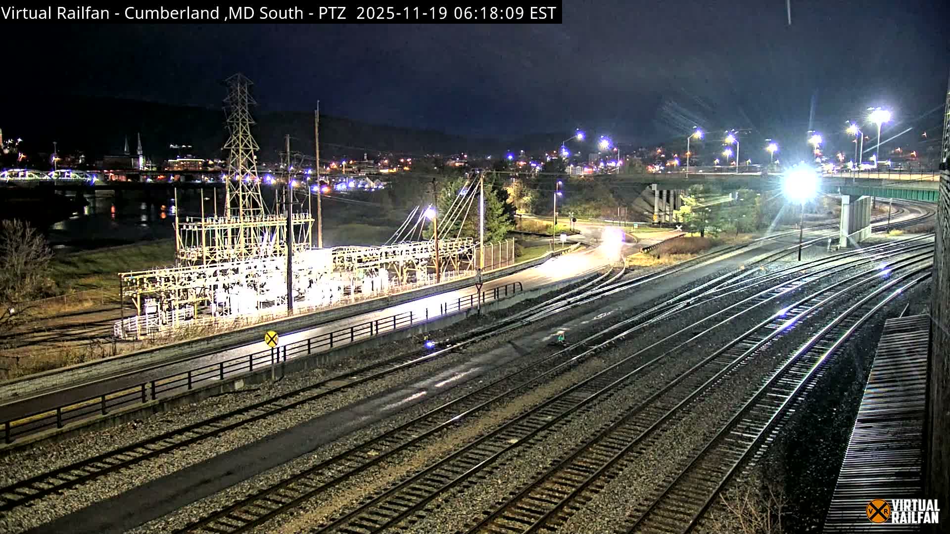A nighttime view of railroad tracks intersecting with roads and a power substation, under clear skies.