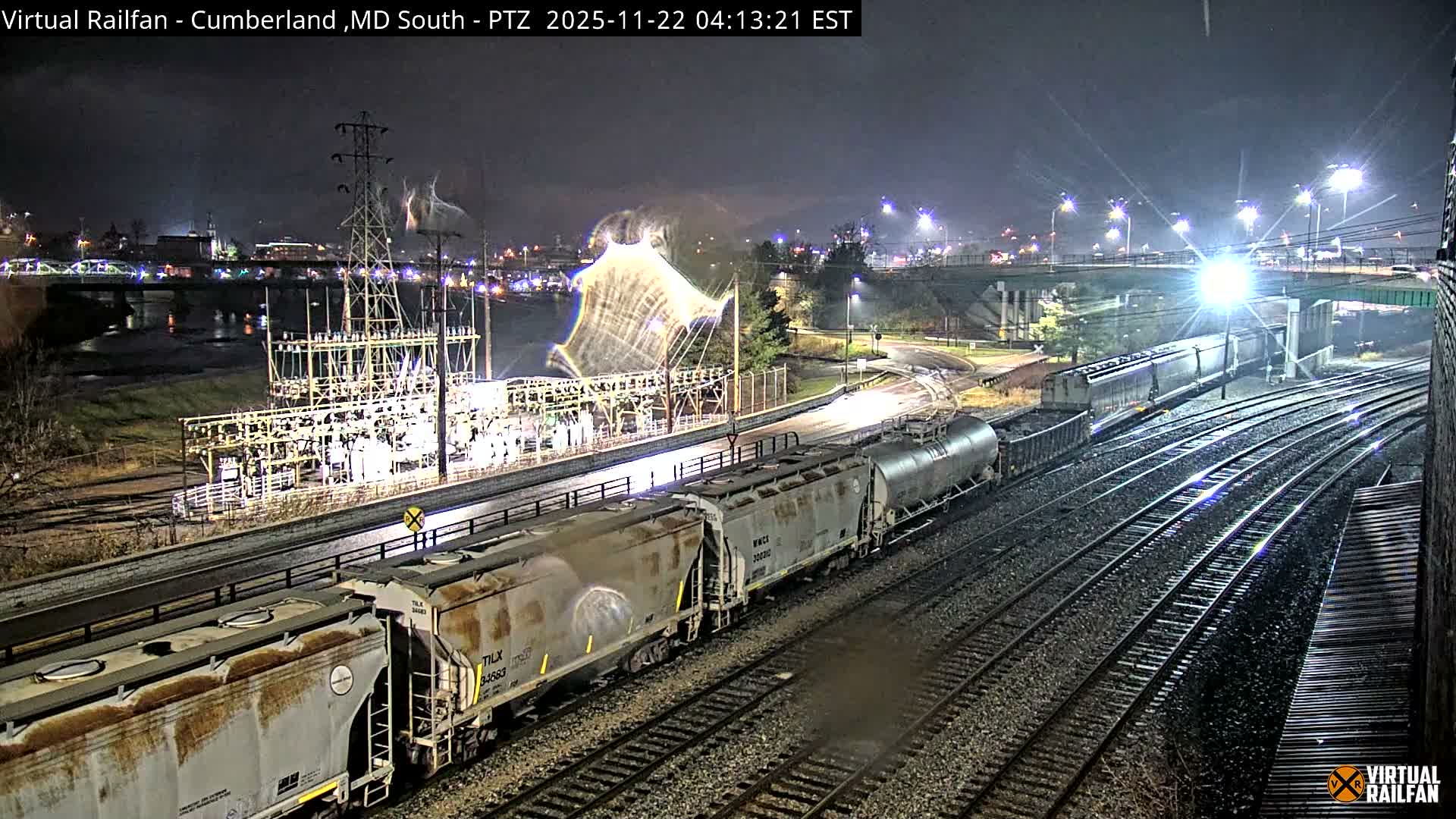 A long freight train rests on multiple tracks at night, set against a glowing power substation, distant city lights, and bridges under a clear sky.