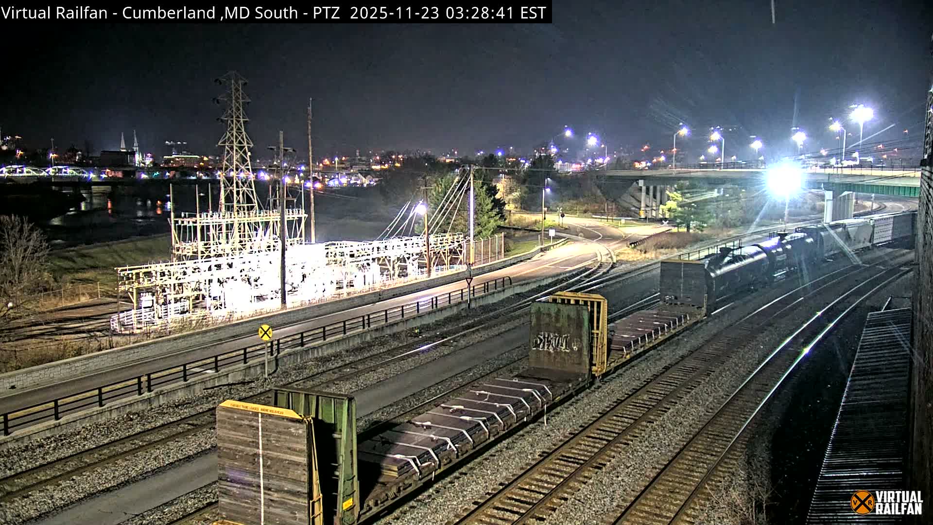 A clear night reveals a train with a bright headlight and multiple freight cars on active tracks, surrounded by an illuminated industrial structure, a distant river with a lit bridge, and city lights under an overpass.