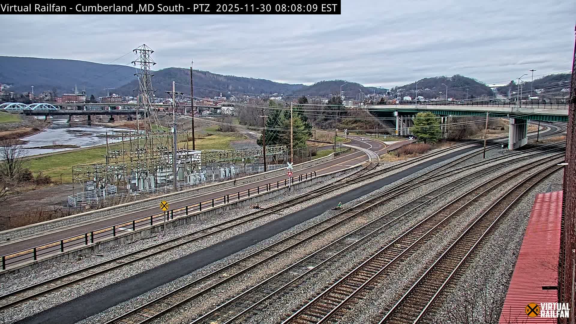 A clear night reveals a train with a bright headlight and multiple freight cars on active tracks, surrounded by an illuminated industrial structure, a distant river with a lit bridge, and city lights under an overpass.