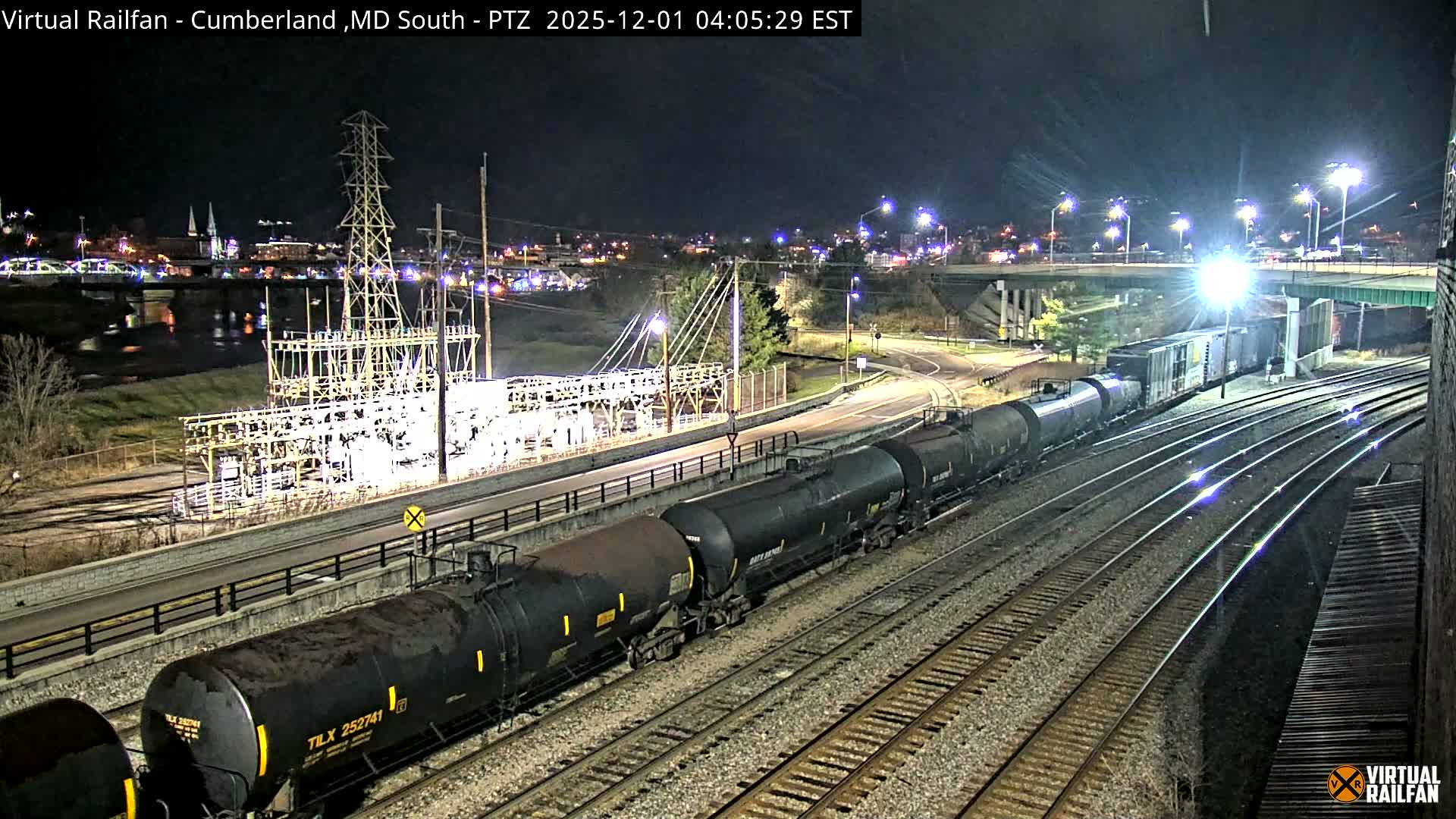 A long freight train with black tank cars moves along multiple tracks at night, adjacent to a brightly lit electrical substation and below an illuminated road bridge, with a city skyline visible across water in the background under a clear sky.
