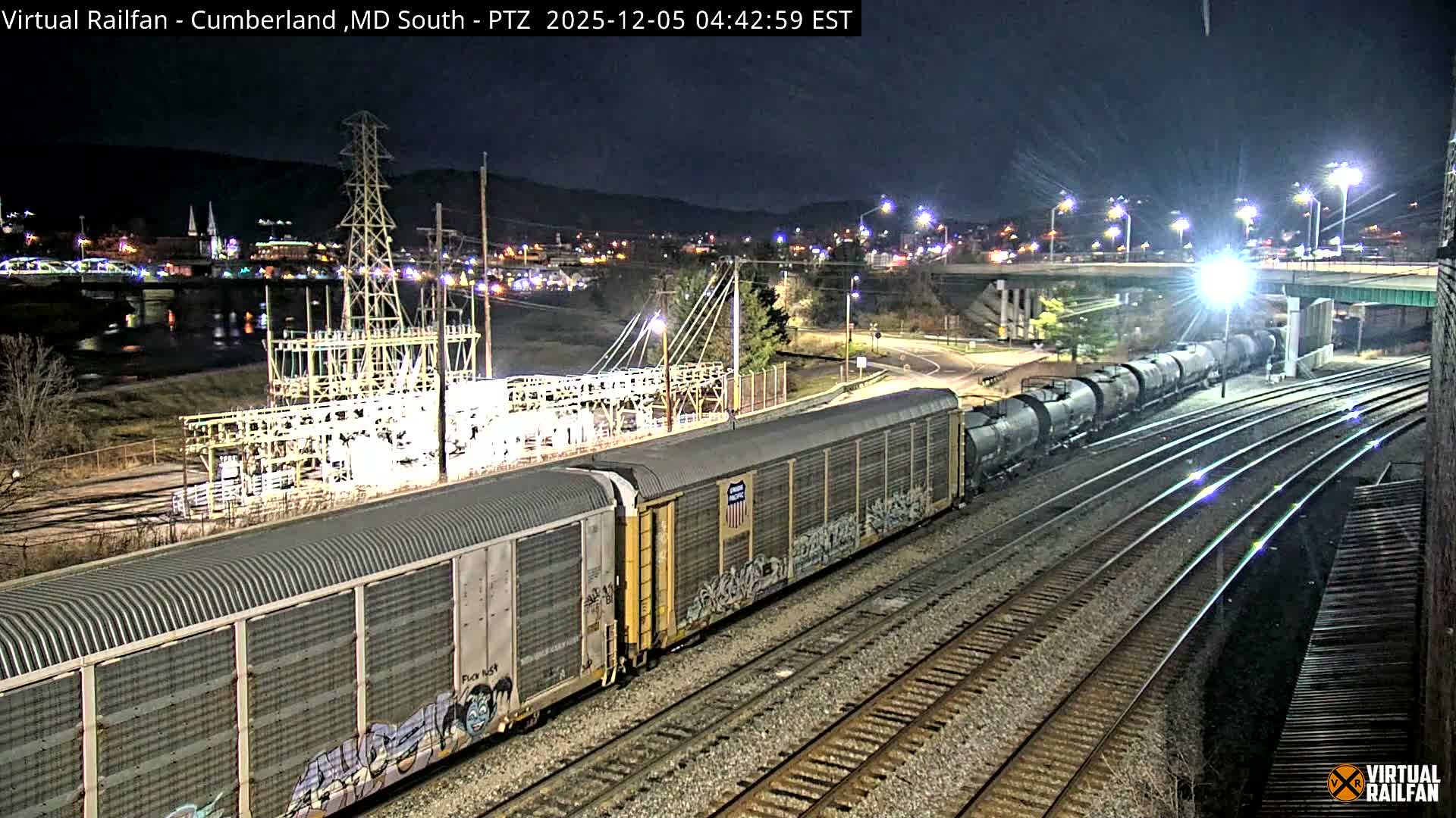 A long freight train, consisting of both boxcars and tank cars, is positioned on multiple tracks in an industrial area at night under a clear sky, with a brightly lit power substation, an overhead bridge, and distant city lights illuminating the dark landscape.
