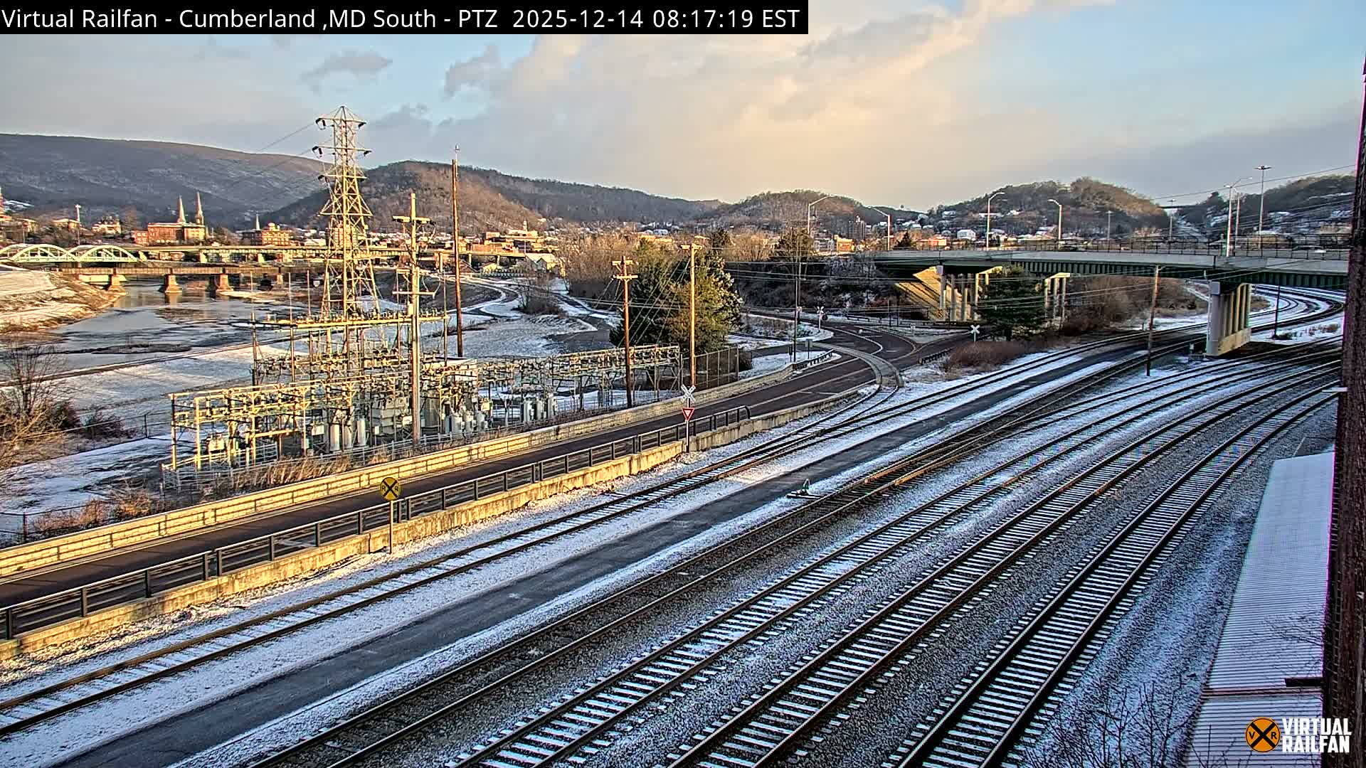 A long freight train, consisting of both boxcars and tank cars, is positioned on multiple tracks in an industrial area at night under a clear sky, with a brightly lit power substation, an overhead bridge, and distant city lights illuminating the dark landscape.