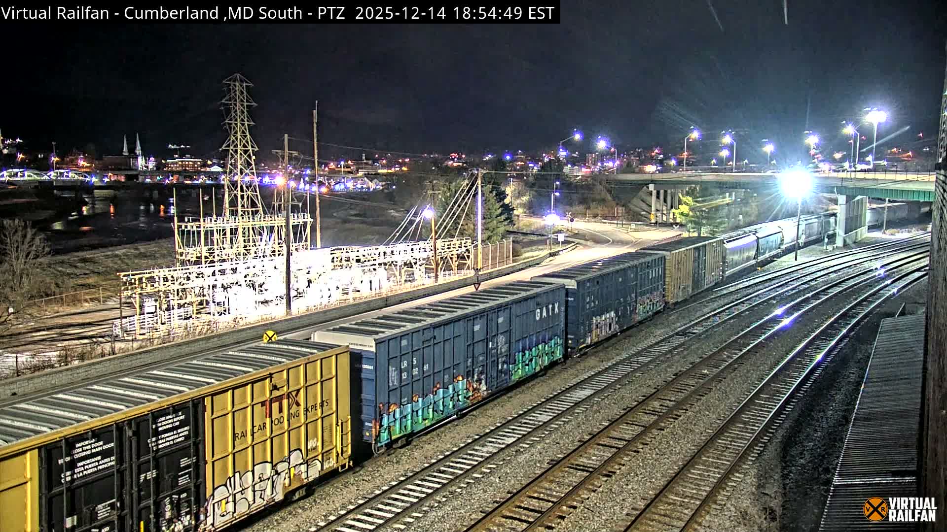 A long freight train, consisting of both boxcars and tank cars, is positioned on multiple tracks in an industrial area at night under a clear sky, with a brightly lit power substation, an overhead bridge, and distant city lights illuminating the dark landscape.