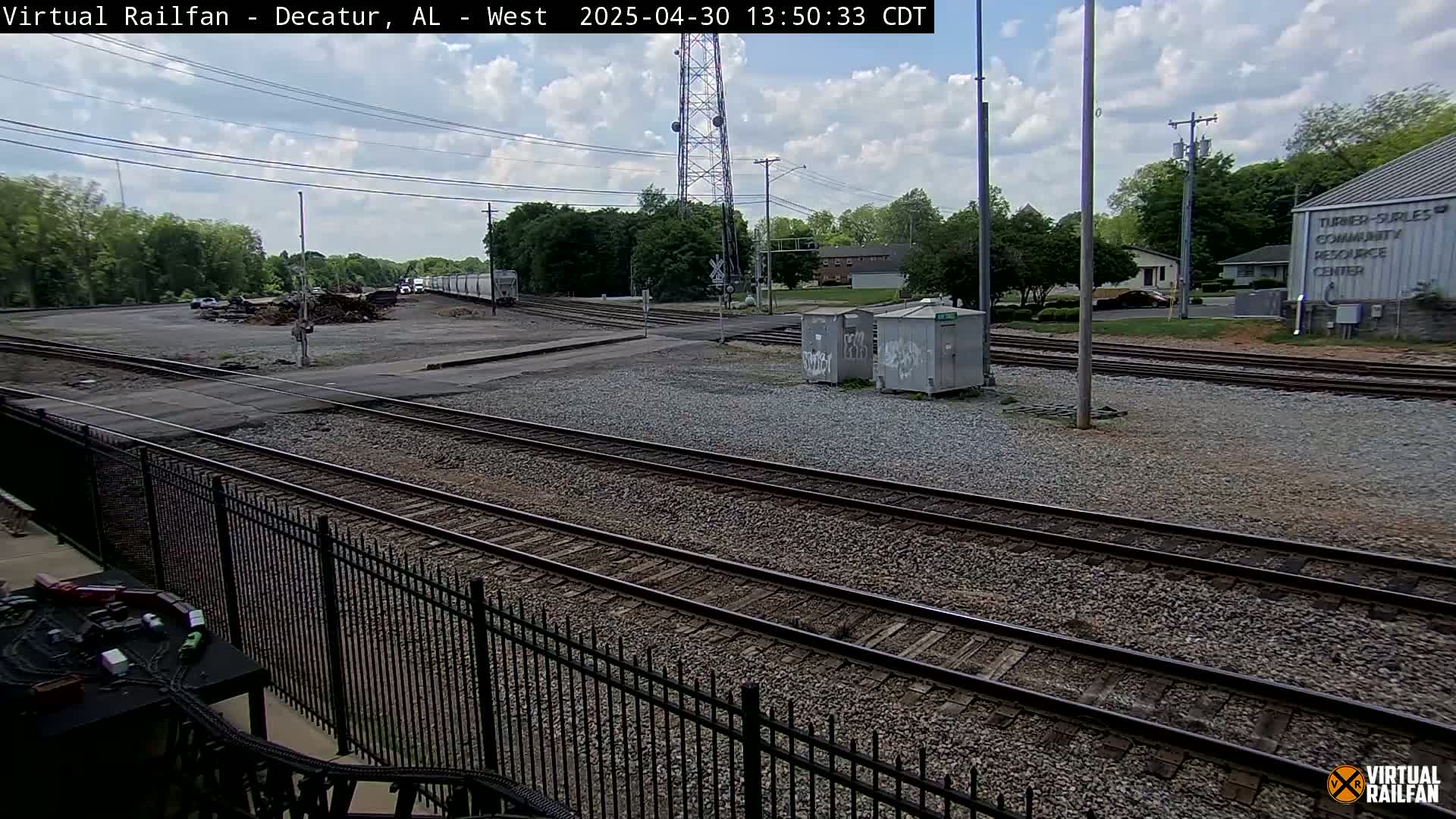 A partly cloudy day reveals a railway switching yard with multiple tracks, a metal fence, and a community center in the background.