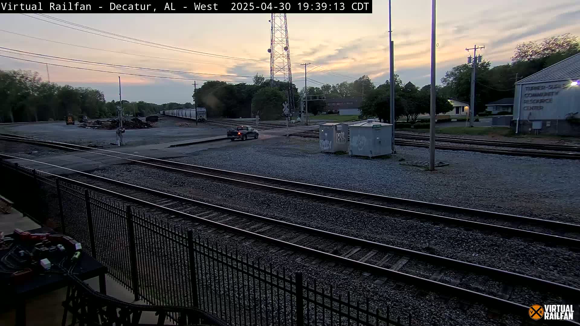 A dusk-lit view of railroad tracks, a nearby road with a vehicle, and some buildings in the background.