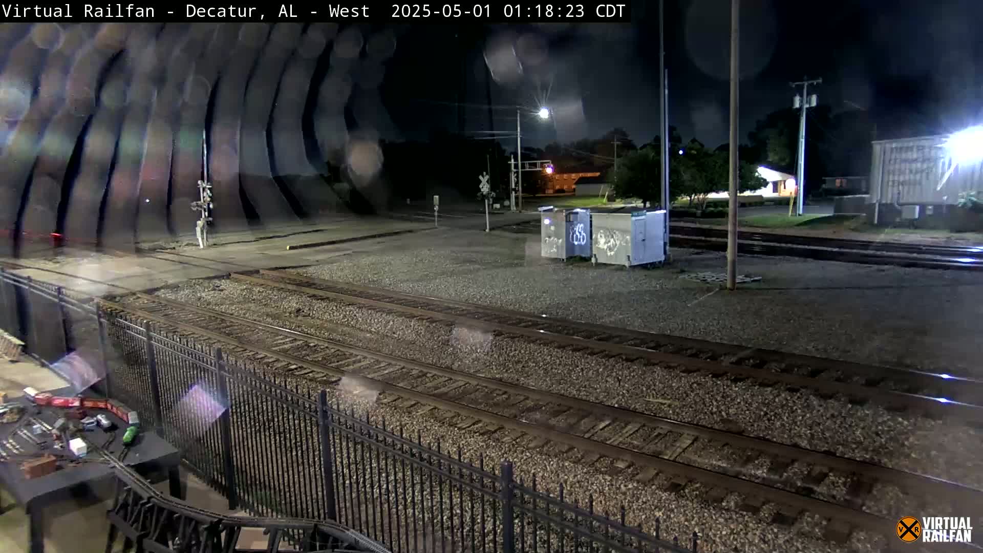 A nighttime view of railroad tracks, a fence, and some buildings under a slightly rainy sky.