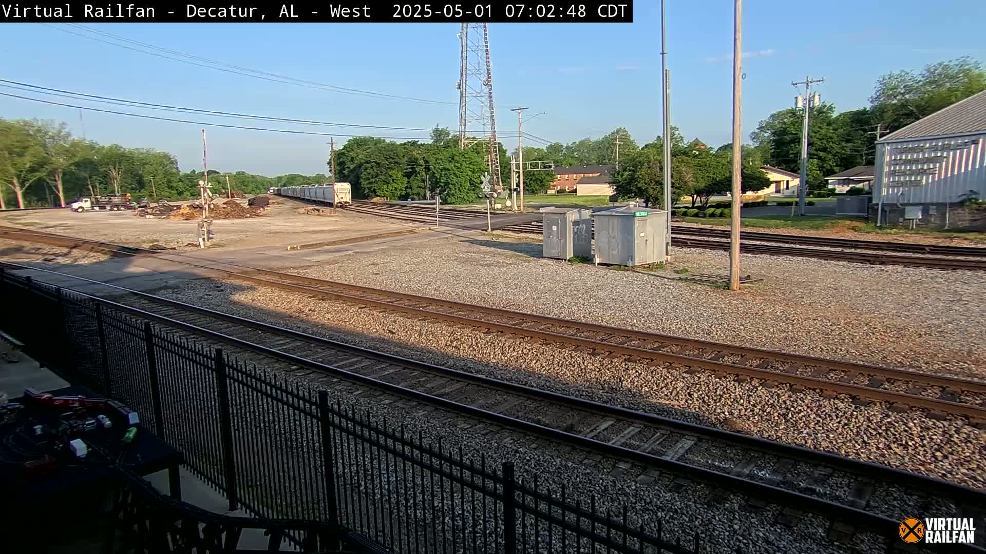A sunny view of a railway yard with multiple train tracks, a gravel surface, some buildings in the background, and trees.