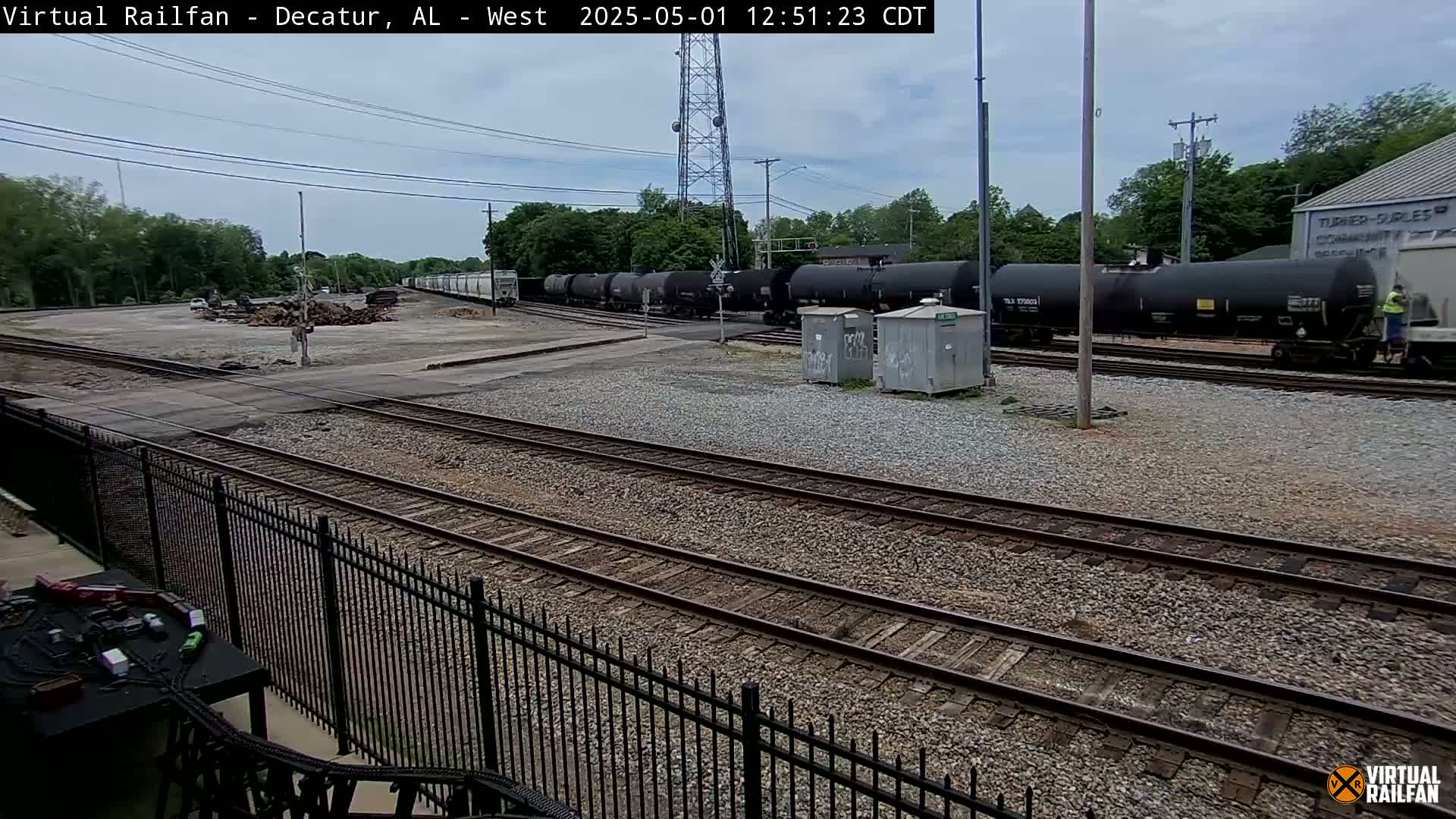A long freight train of black tank cars sits on a railway in a gravelly yard under an overcast sky.